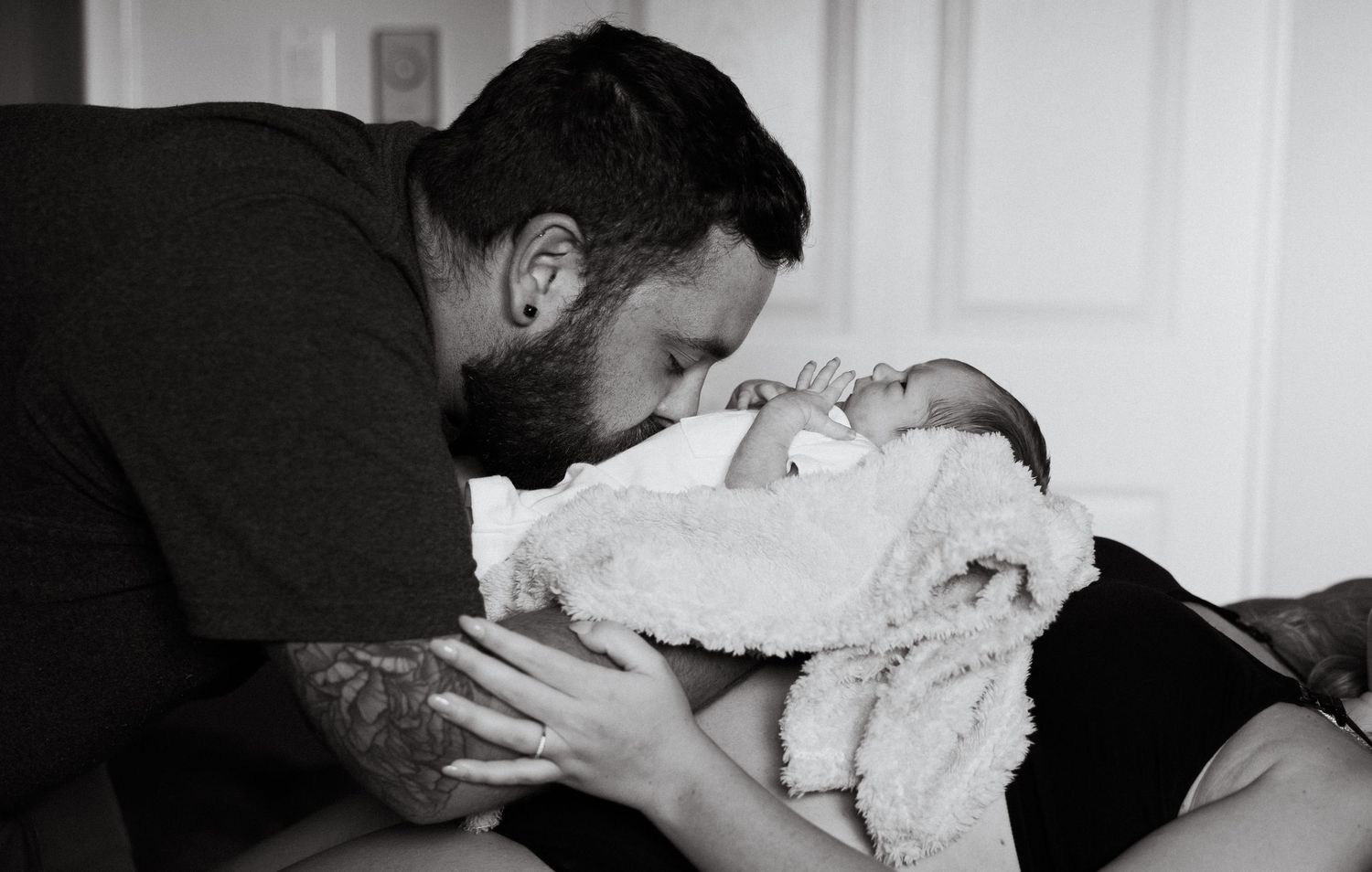 Black and white photo of tattooed father kissing swaddled newborn in hospital.