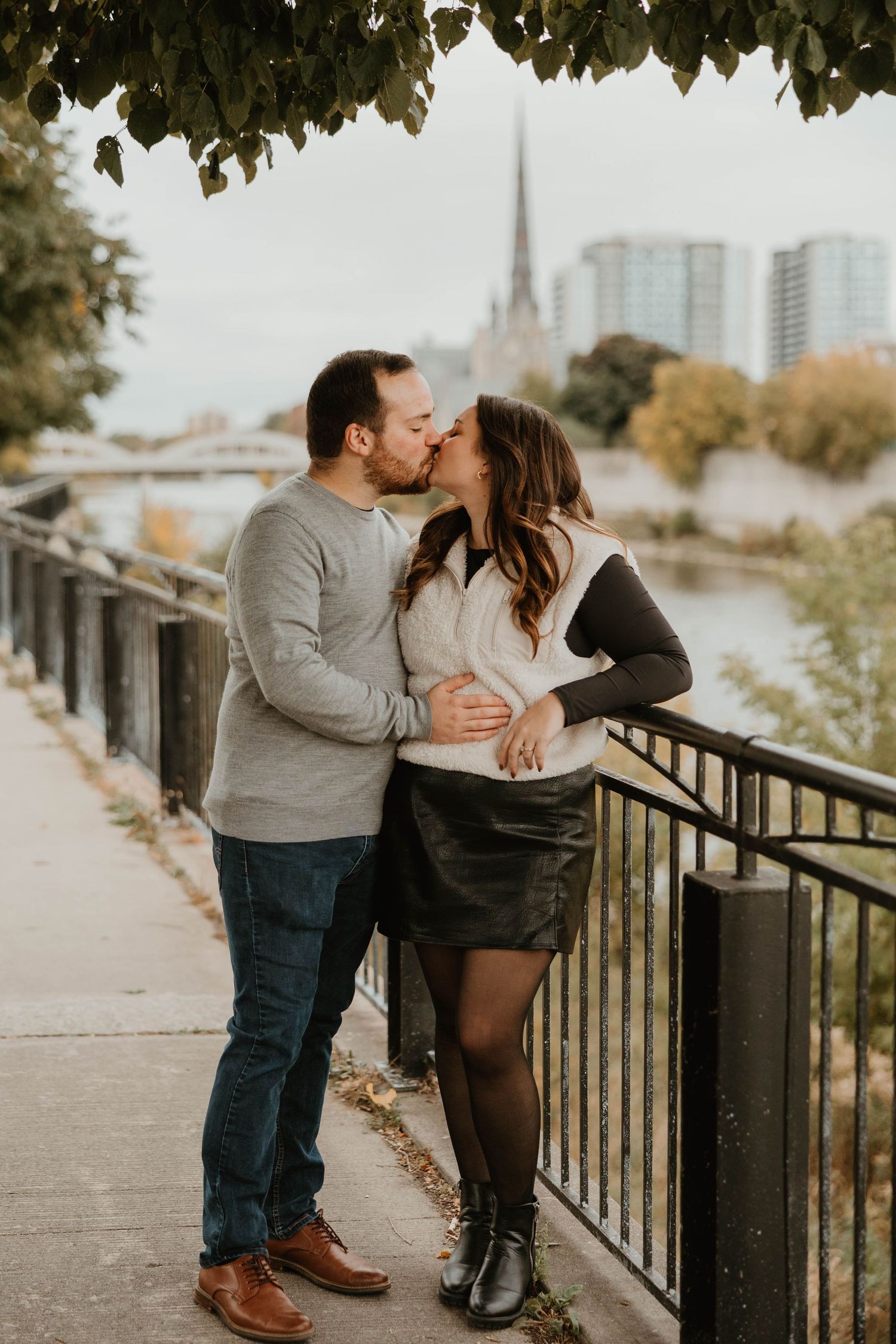 Couple kissing by iron fence along river with city skyline in background.
