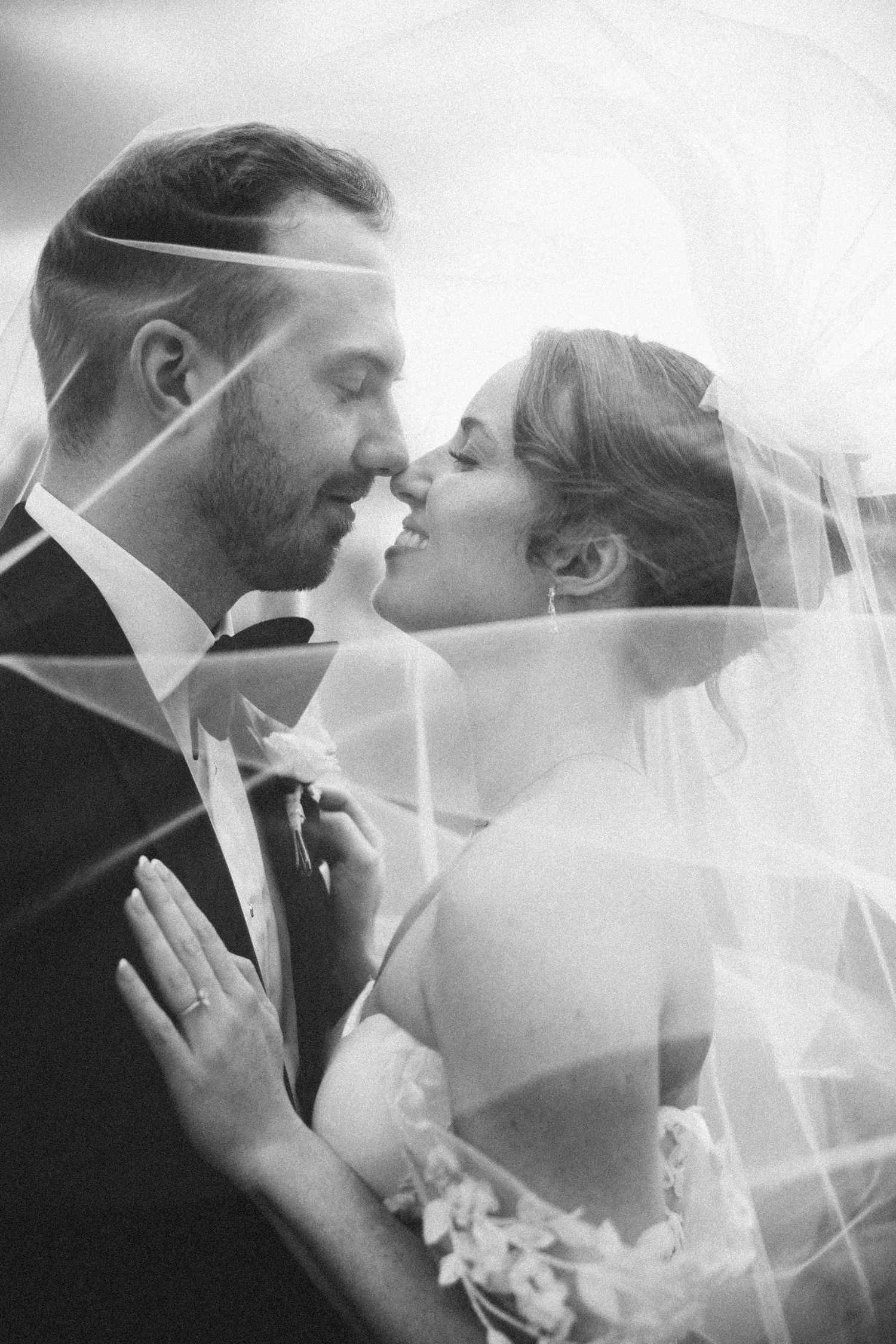 Black and white wedding photo of bride and groom almost kissing under a veil, captured in a romantic and intimate moment.