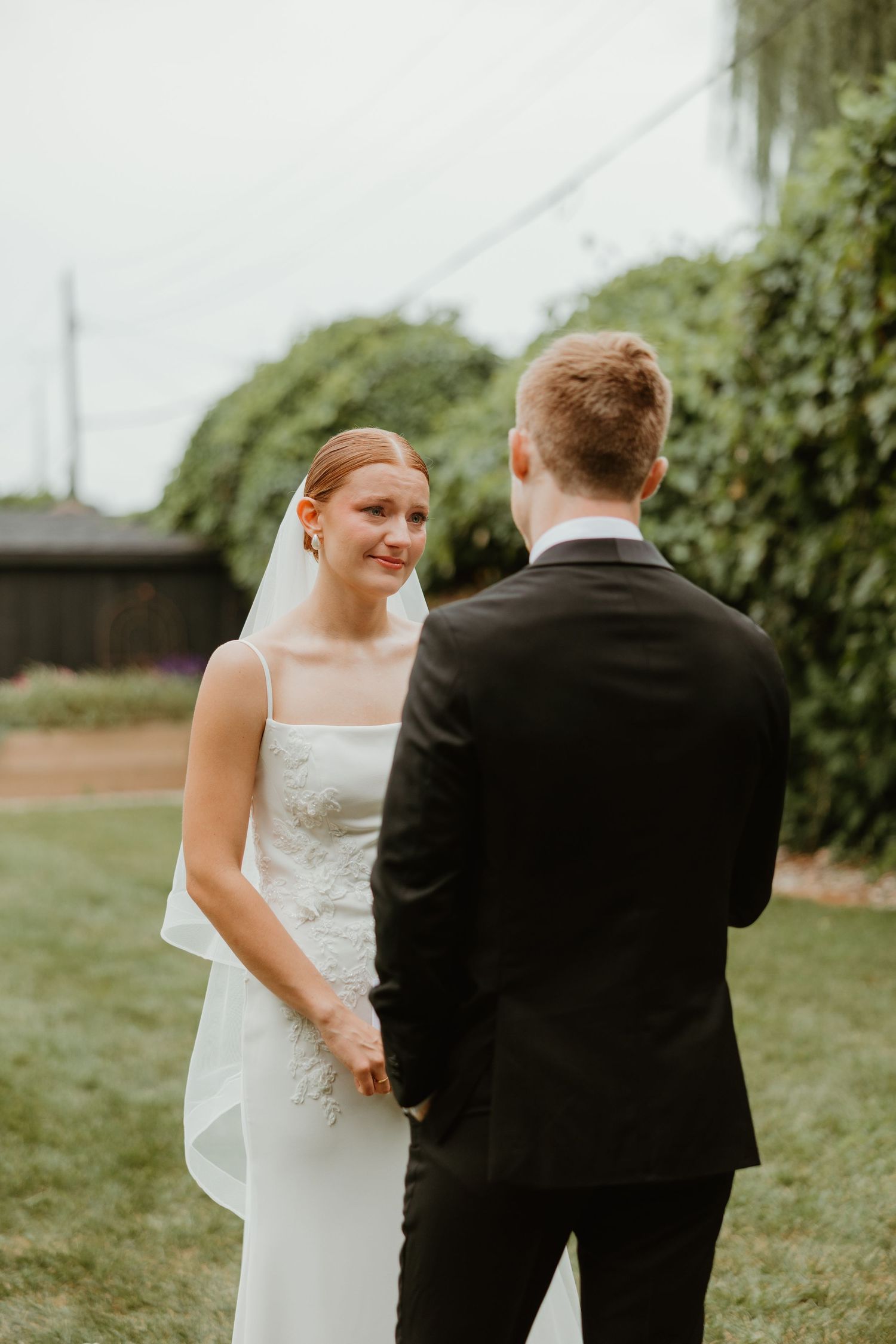 Bride in white lace gown holds grooms hands during emotional outdoor first look ceremony.