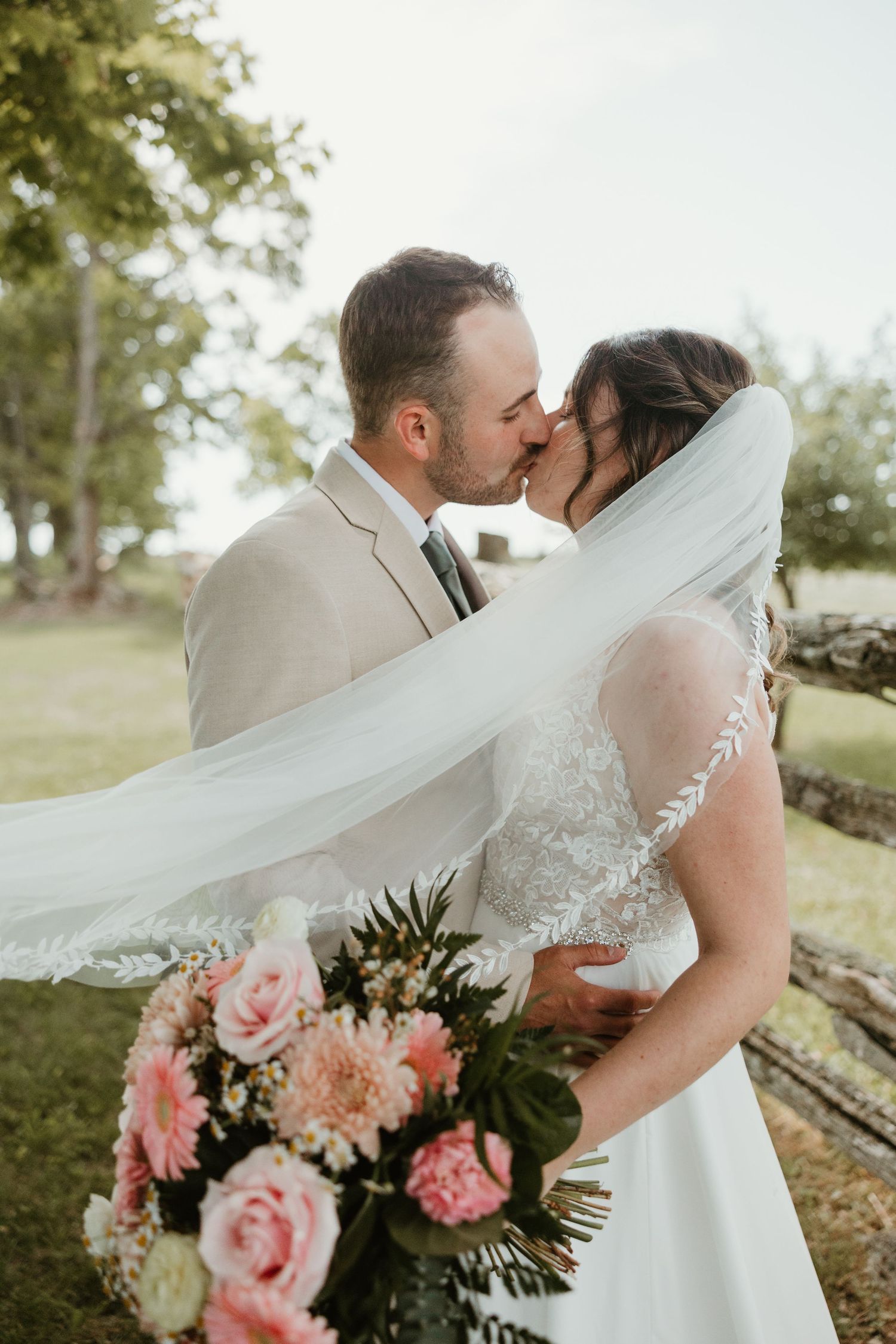 Bride and groom kissing outdoors with flowing lace-trimmed veil and pink floral bouquet.