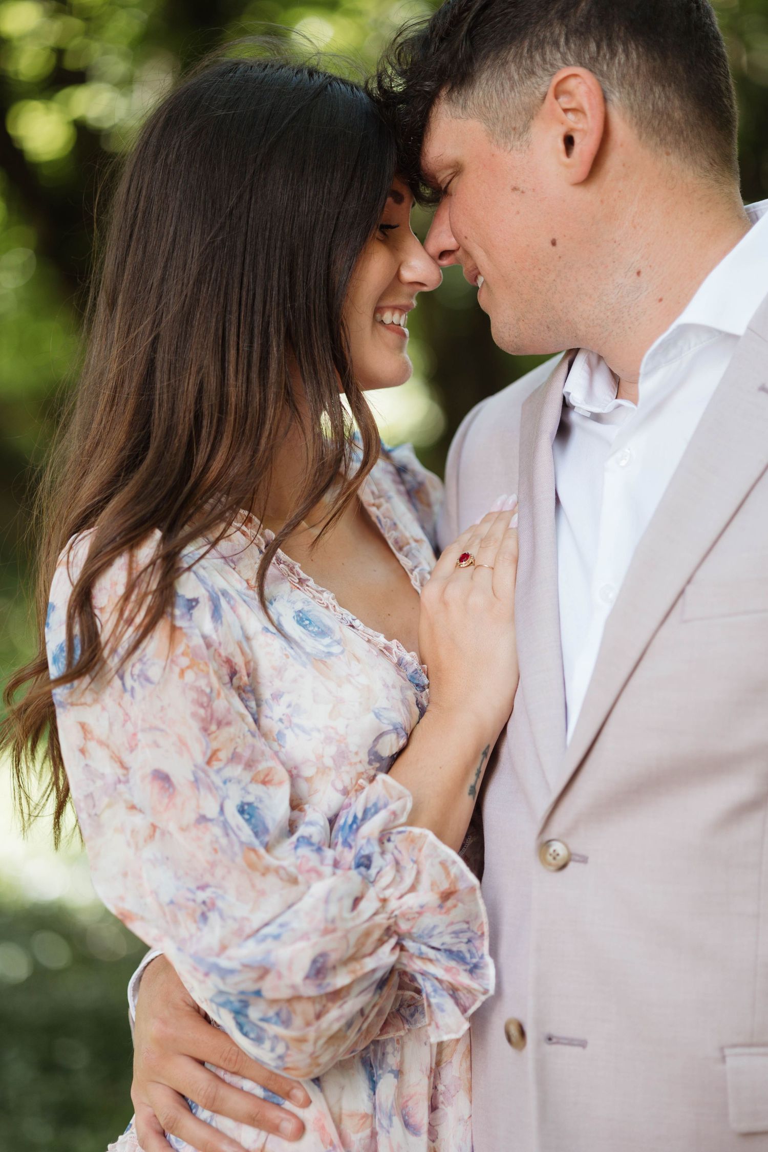 Smiling couple in a floral dress and beige suit embracing closely outdoors in a park.