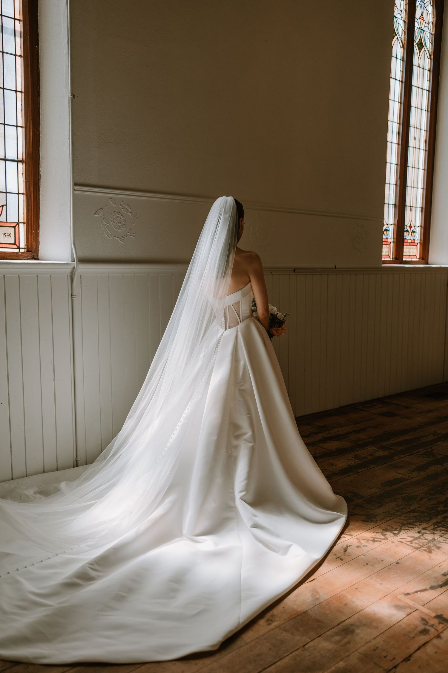 Bride in a stunning white gown with cathedral veil standing before stained glass church windows.