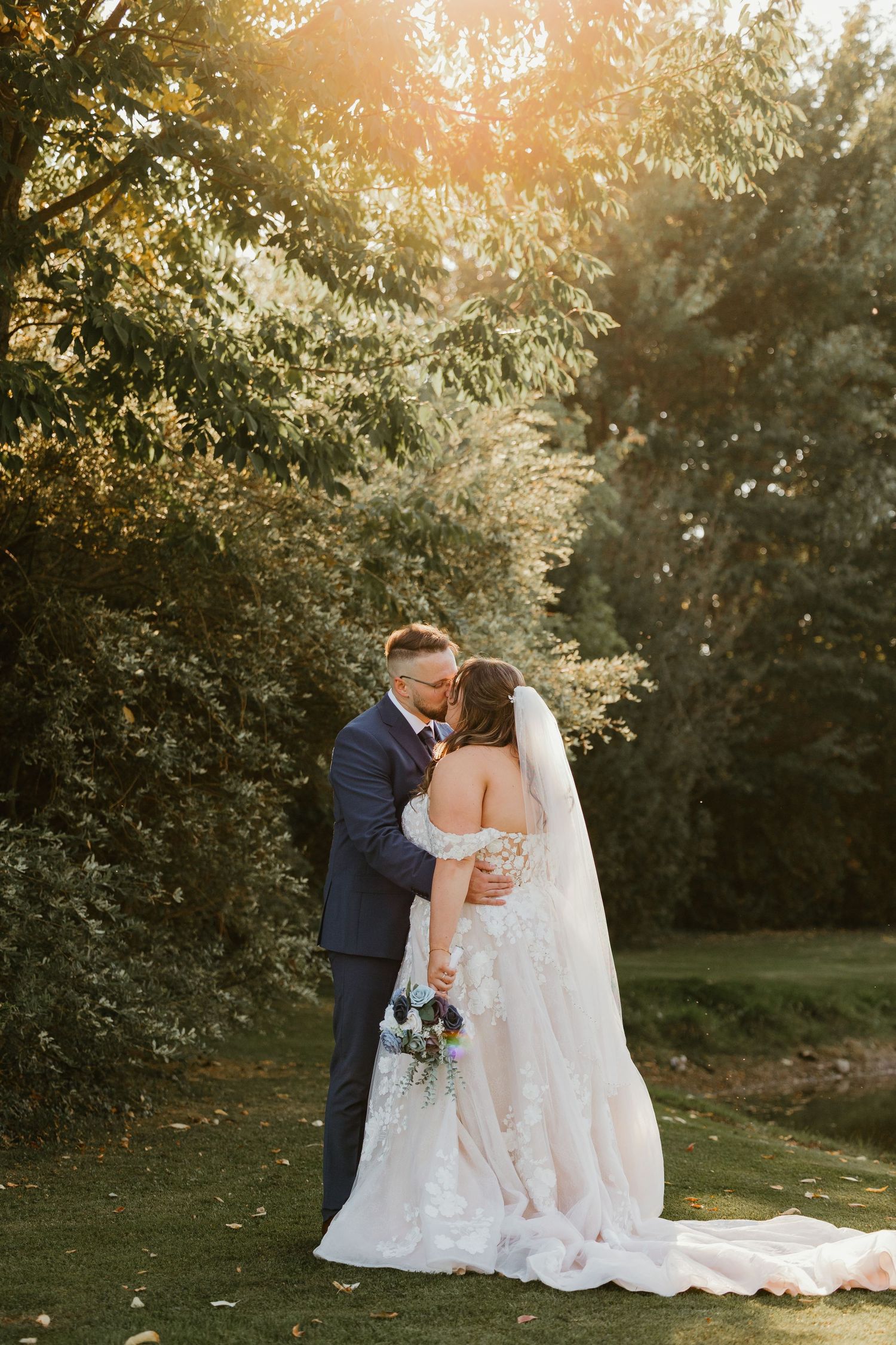 Bride and groom sharing a golden hour kiss outdoors surrounded by lush green trees.
