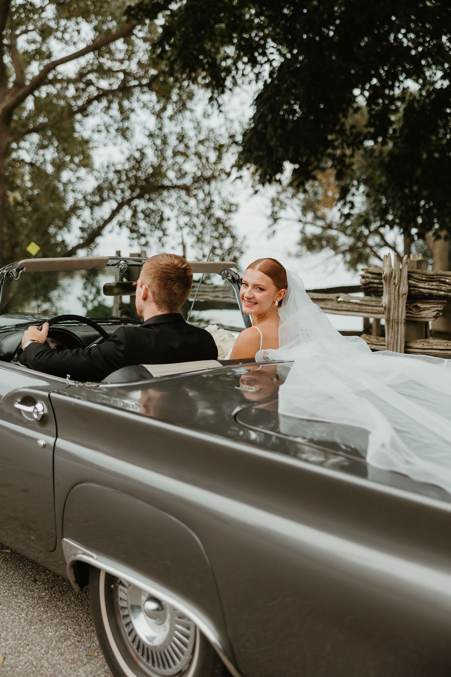 Smiling bride in white veil looks back while riding in a classic gray convertible with groom.
