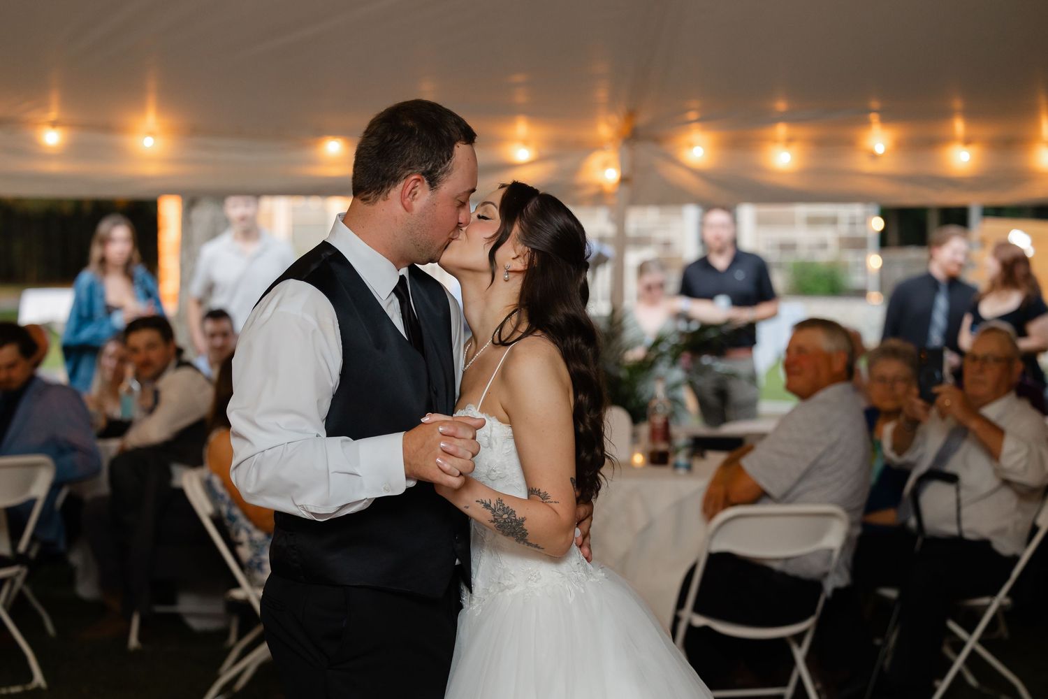 Bride and groom share first dance kiss under string lights at elegant tented wedding reception.