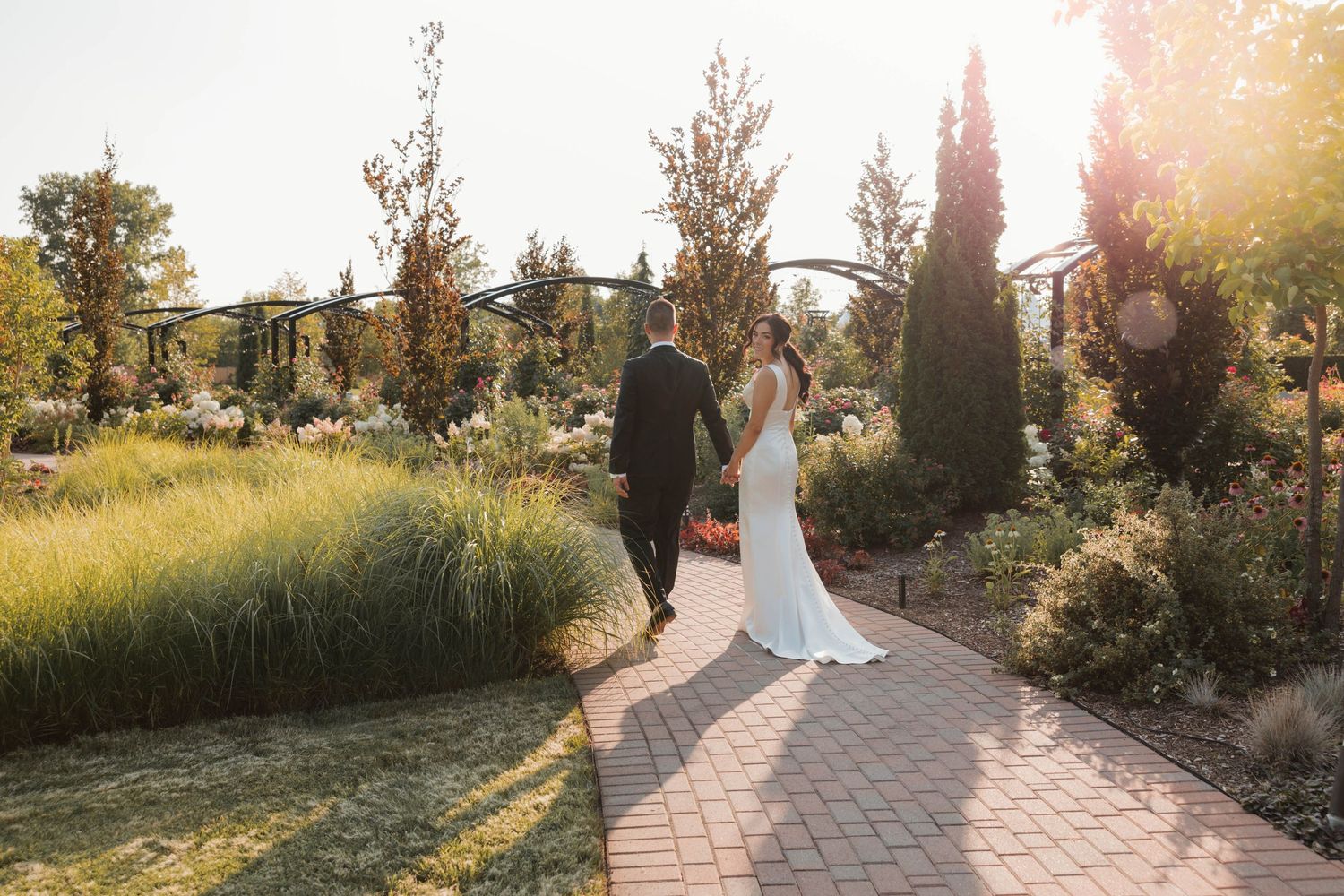 Bride and groom hold hands walking along a brick garden path surrounded by lush greenery in golden sunlight.