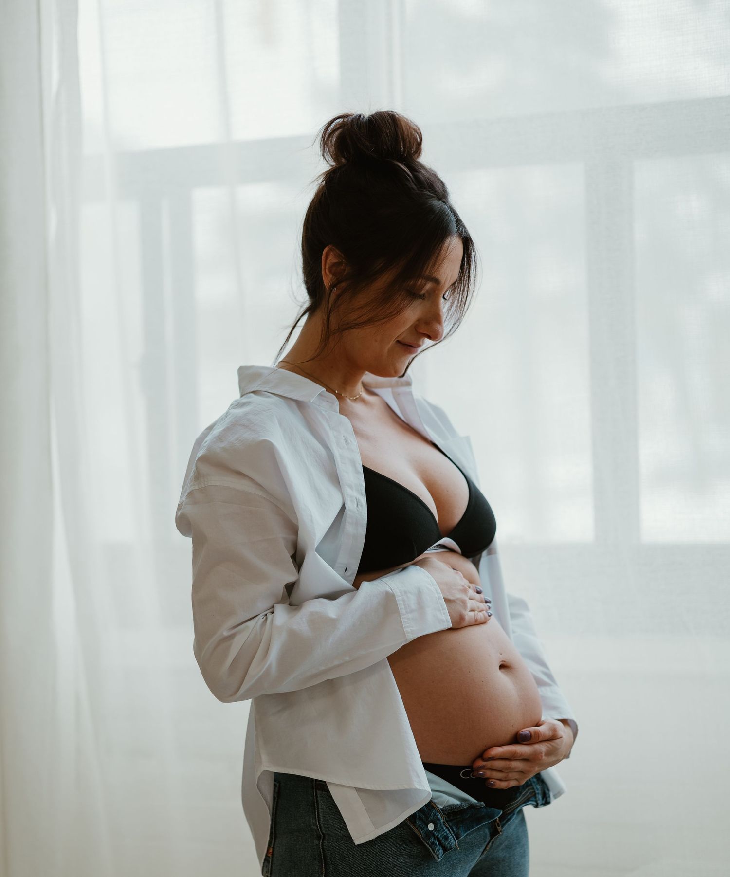 Pregnant woman in black bra and open white shirt cradling her baby bump by sheer curtains.