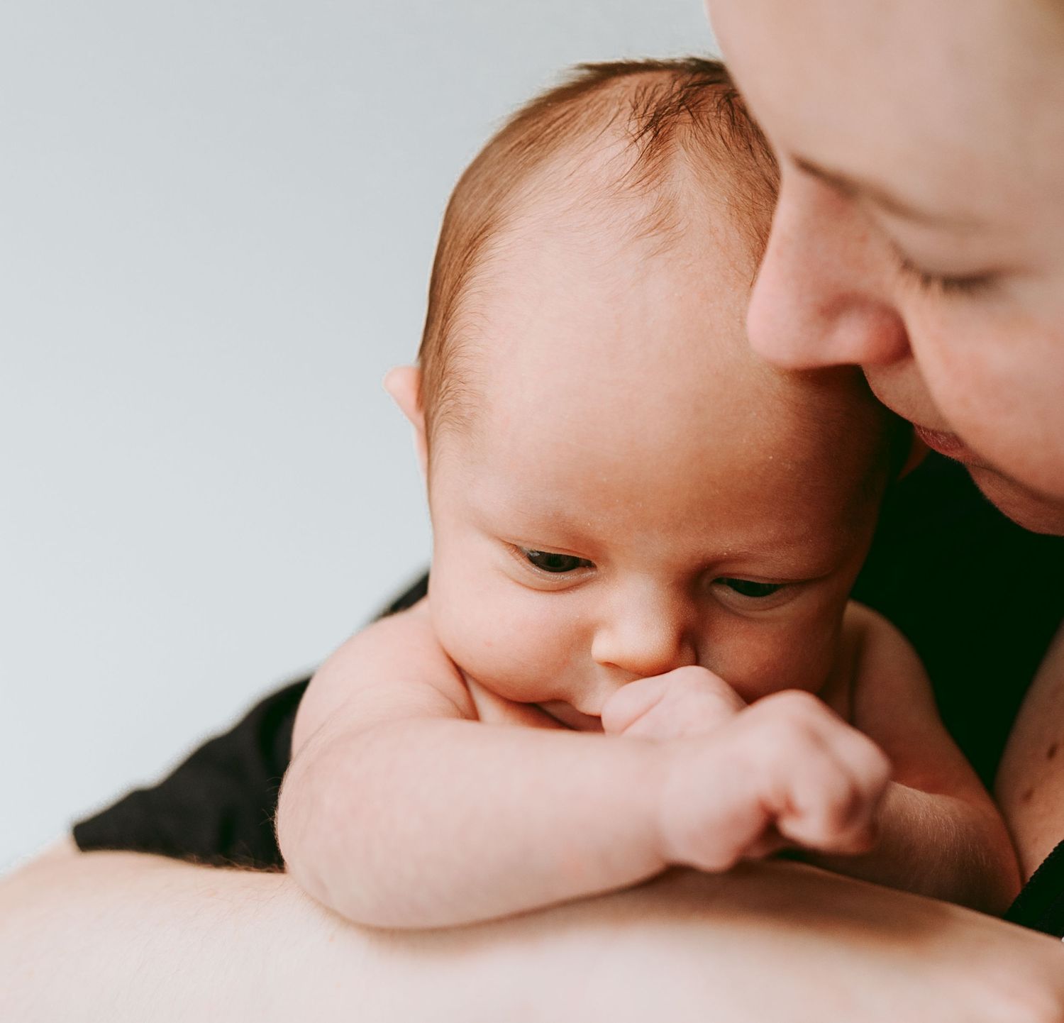 Close-up of parent gazing at newborn baby sucking on tiny fist.