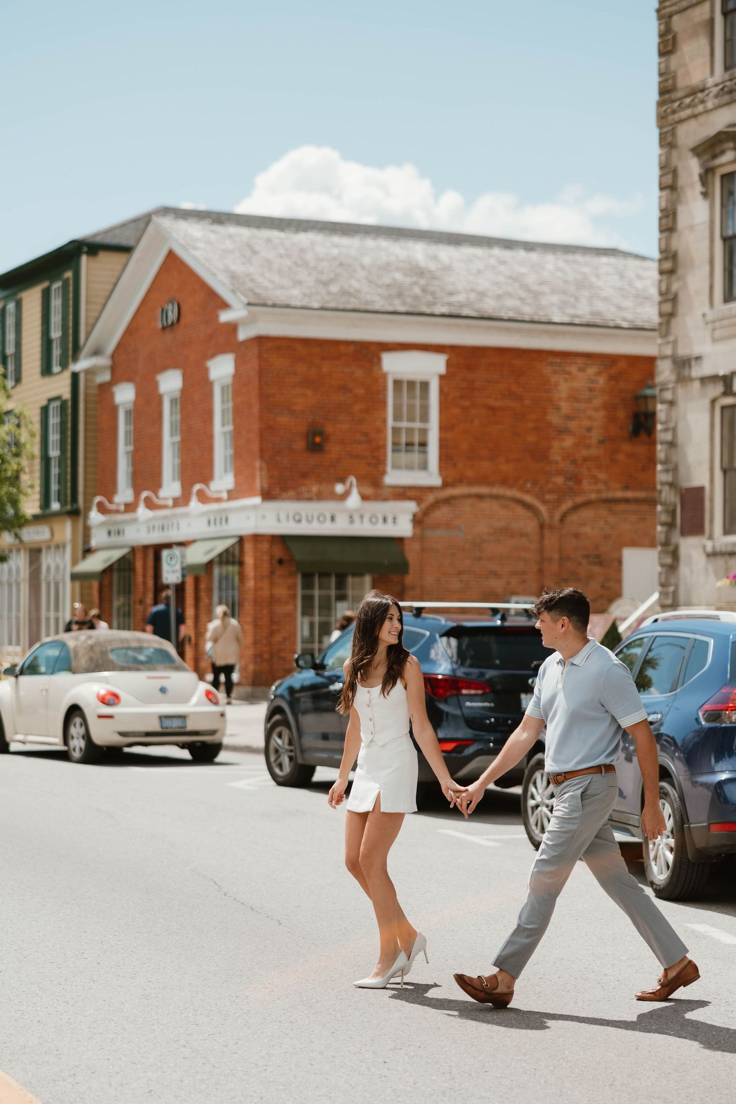 Couple holding hands crossing a sunny small-town street with brick buildings behind them.