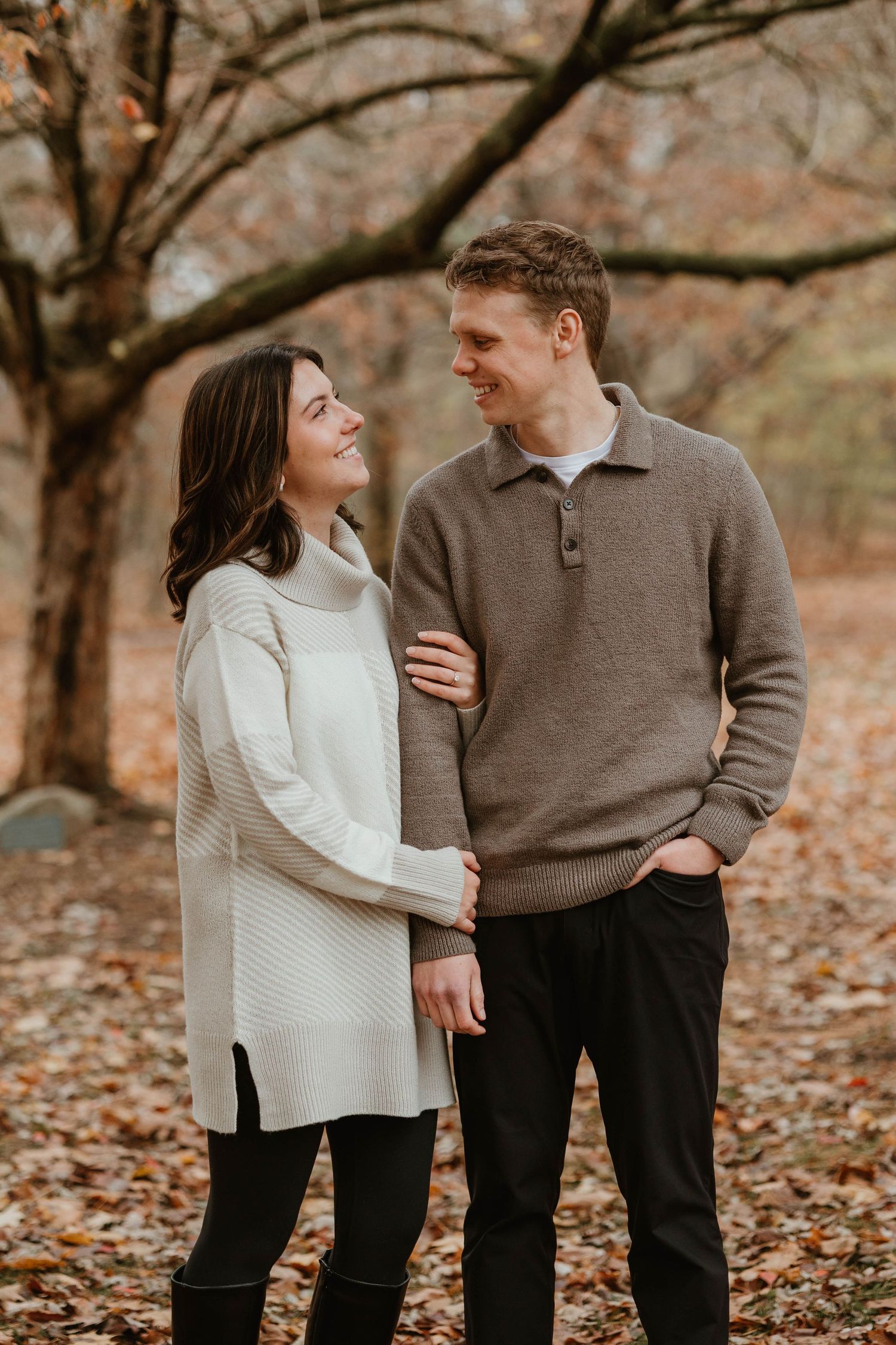 Couple smiling at each other in autumn park, woman in cream sweater, man in brown pullover.