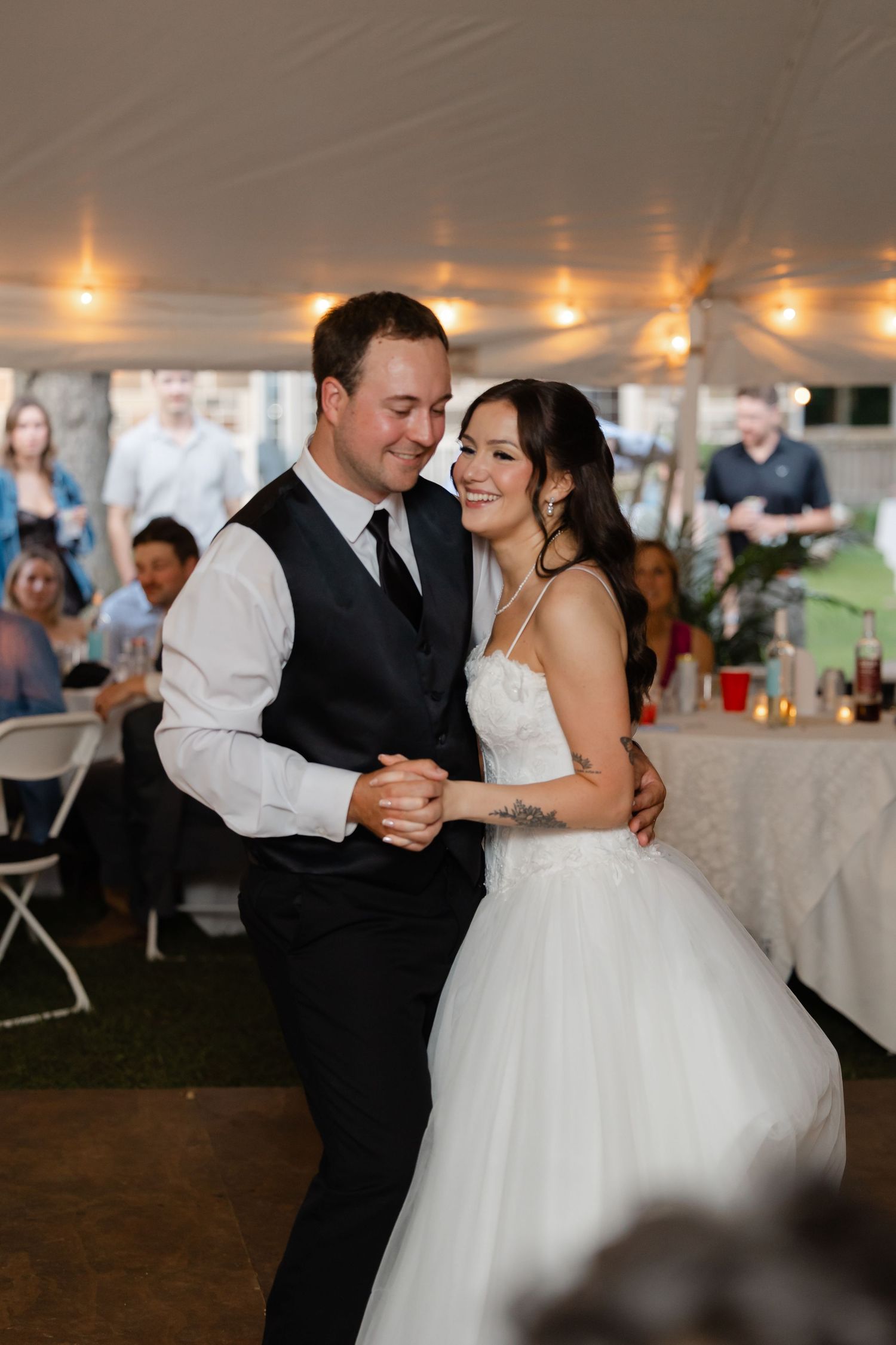 Bride and groom sharing first dance under tent reception, guests watching, string lights glowing.