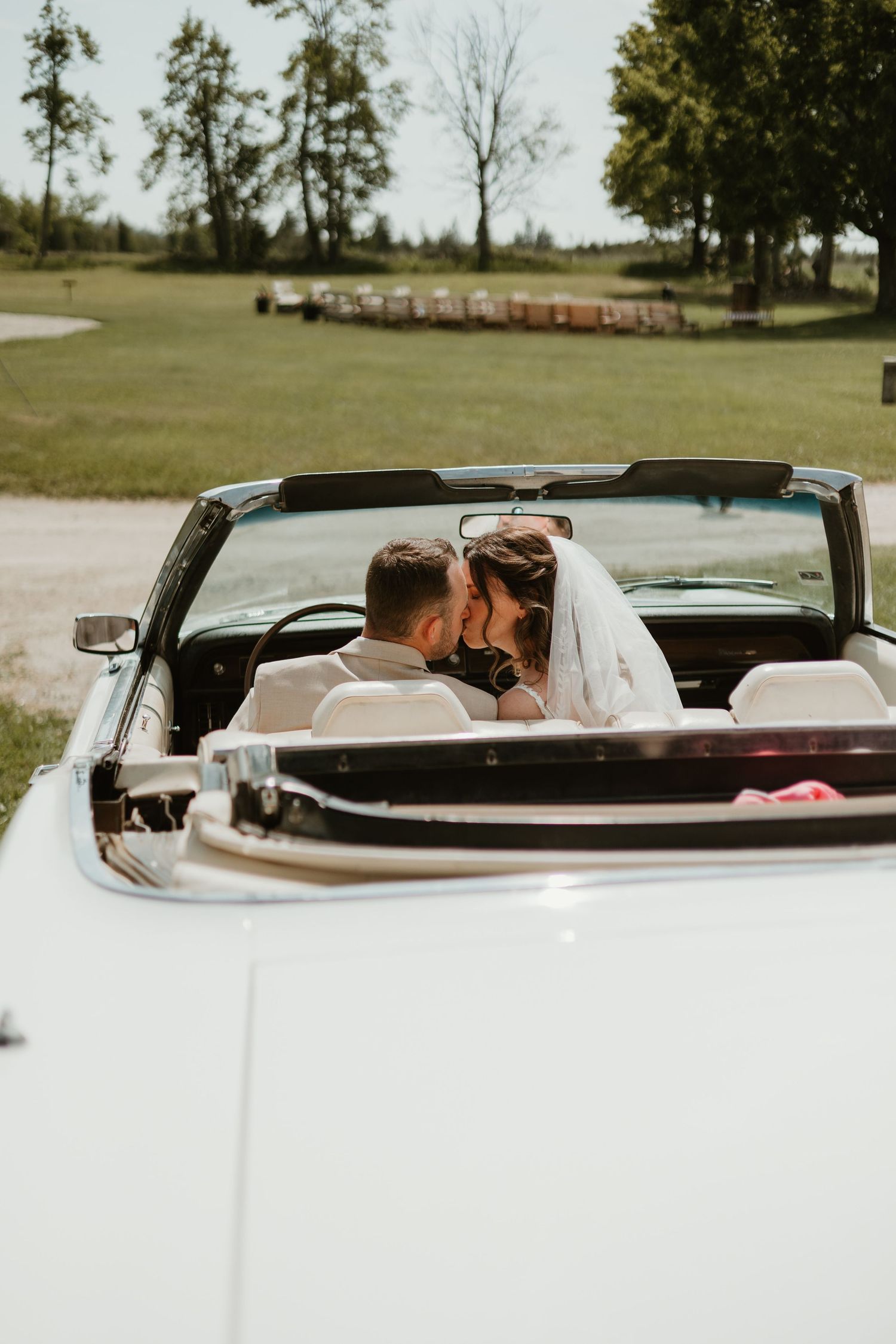 Bride and groom sharing a kiss in a classic white convertible car on their wedding day.