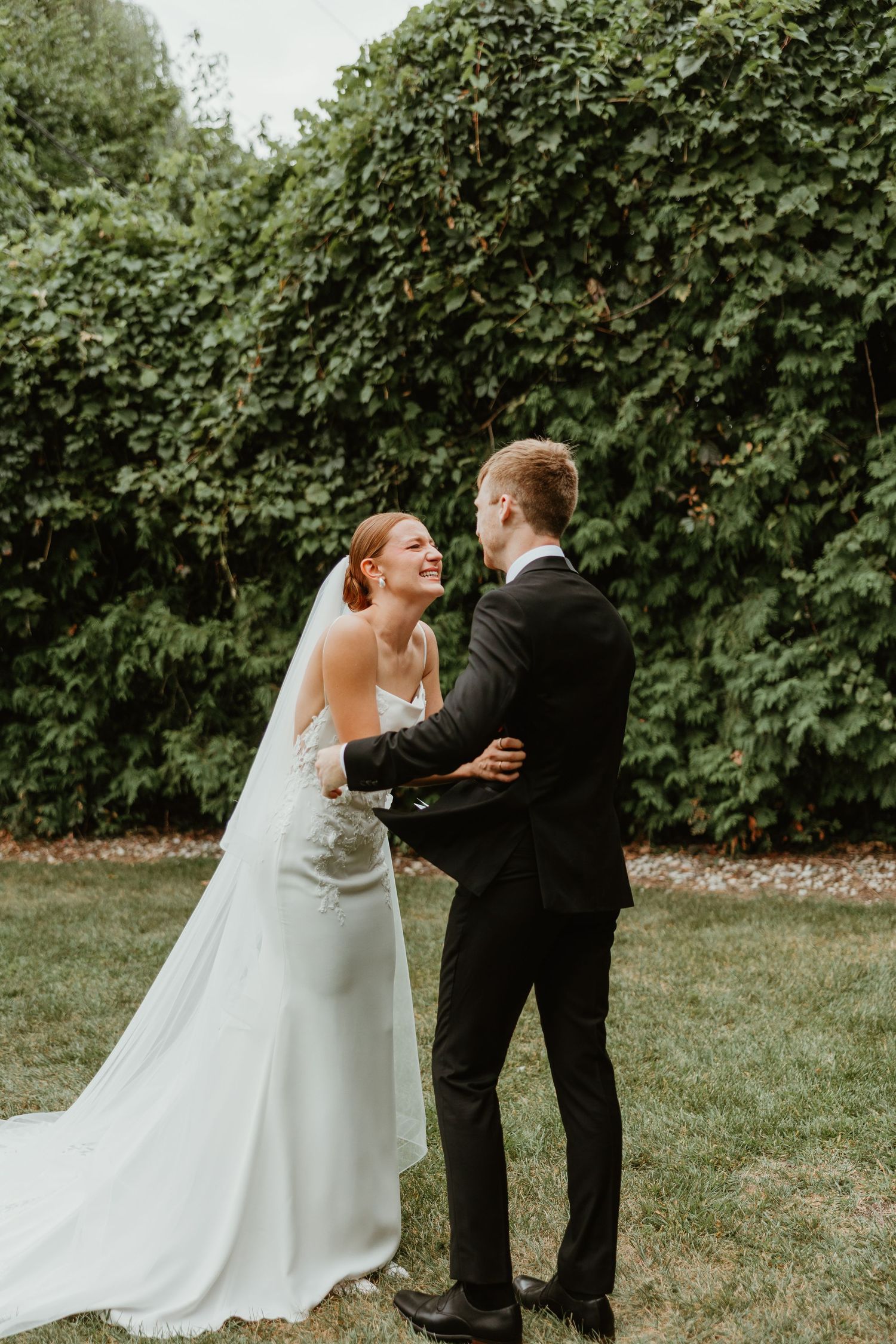 Bride and groom share a joyful moment embracing outdoors with lush green ivy wall backdrop.