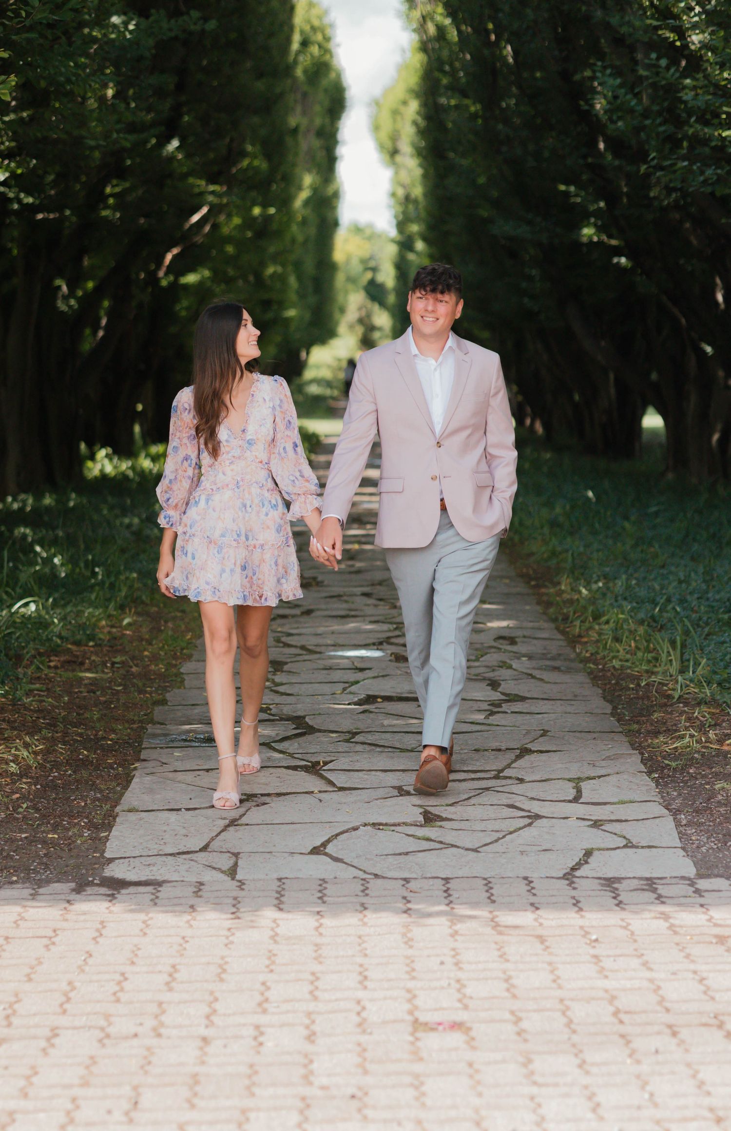 Couple holding hands walking down a tree-lined stone pathway in a lush green park.