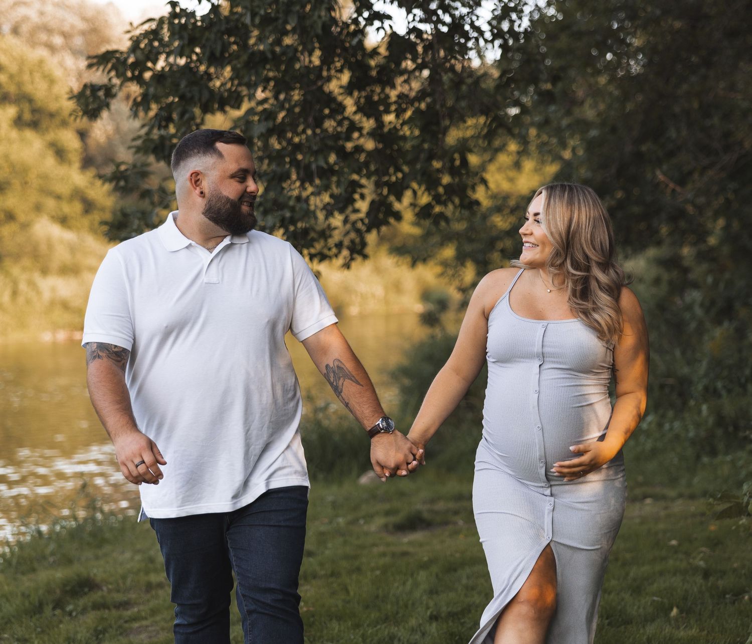 Happy expecting couple holding hands and smiling by a sunny riverside.