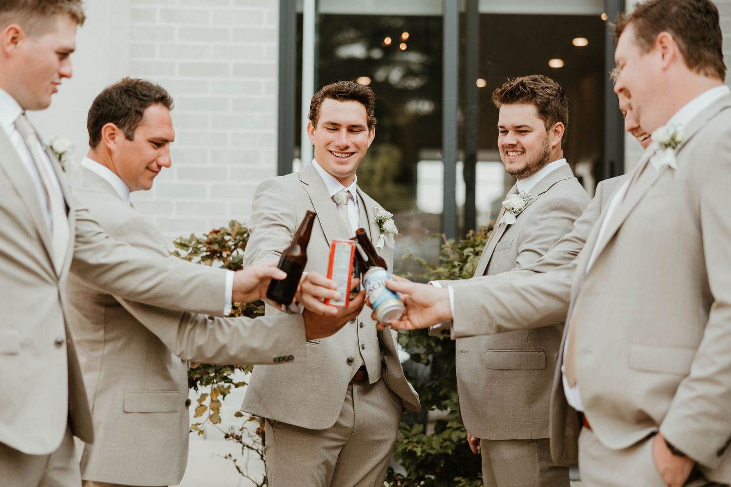 Groomsmen in matching tan suits laugh together while sharing drinks before wedding ceremony.