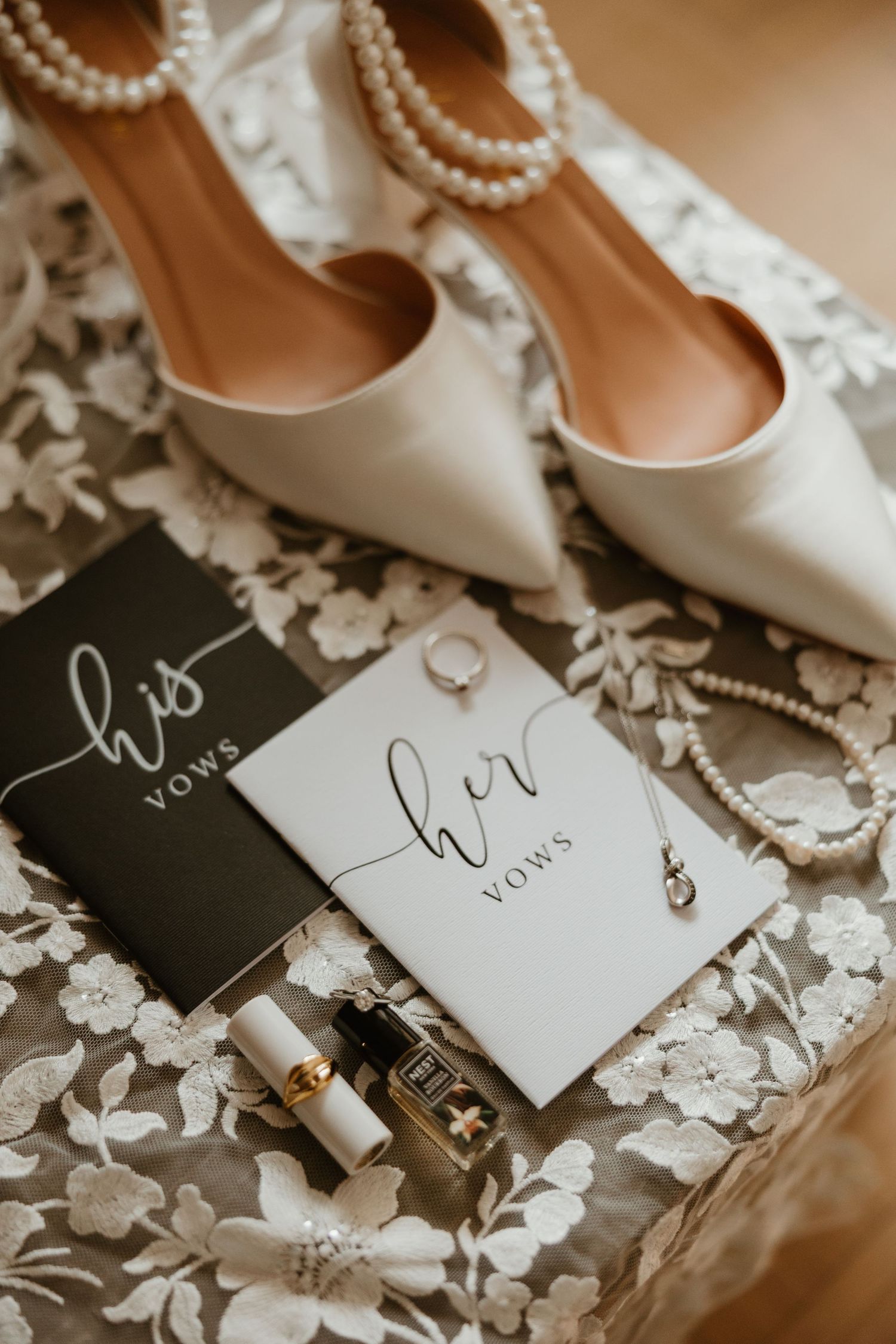 Flat lay of wedding details including his and her vow books, pearl heels, rings, perfume, and jewelry.