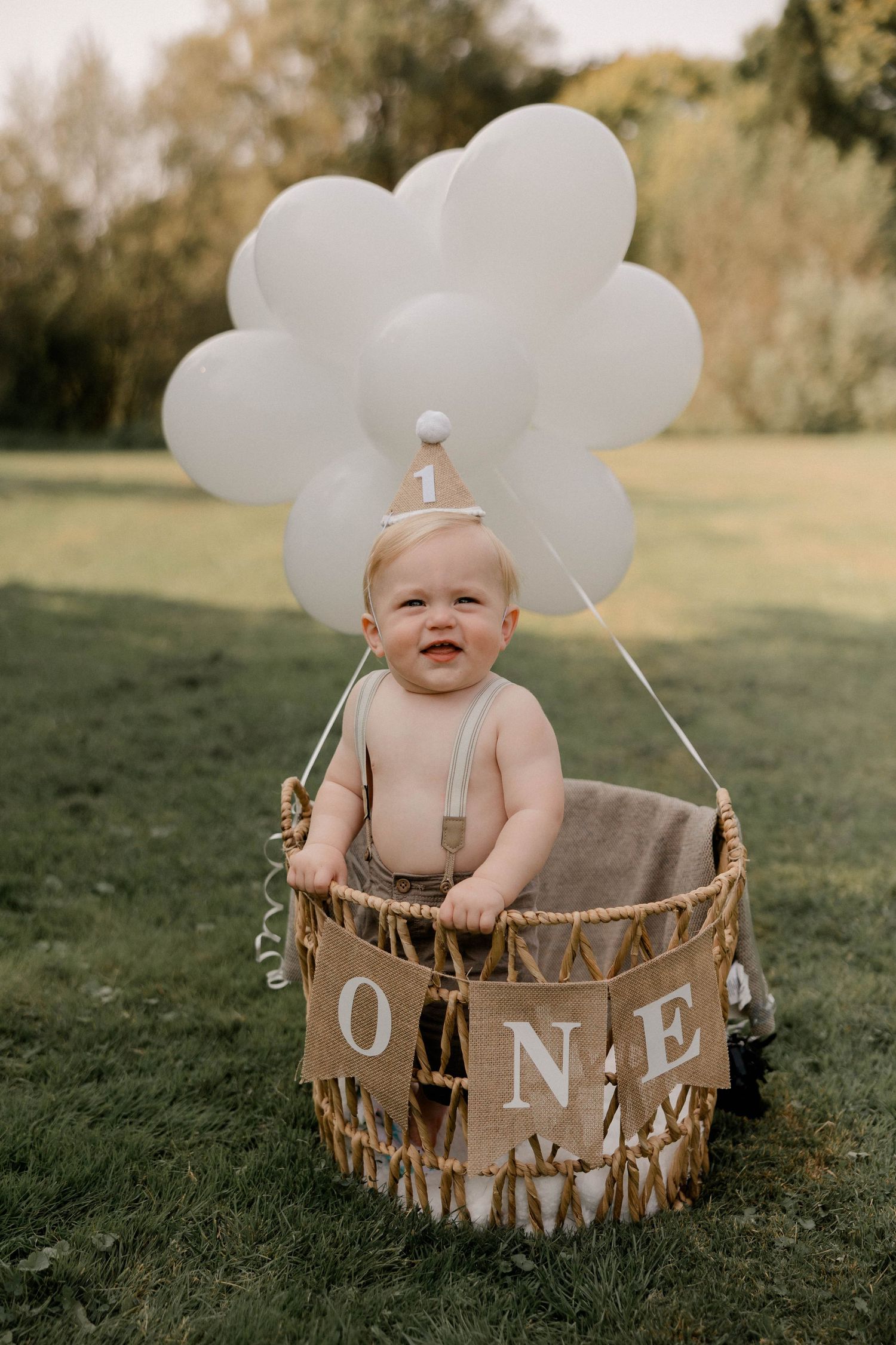 A baby's first birthday photo with child sitting in a basket labeled ONE with white balloons overhead.
