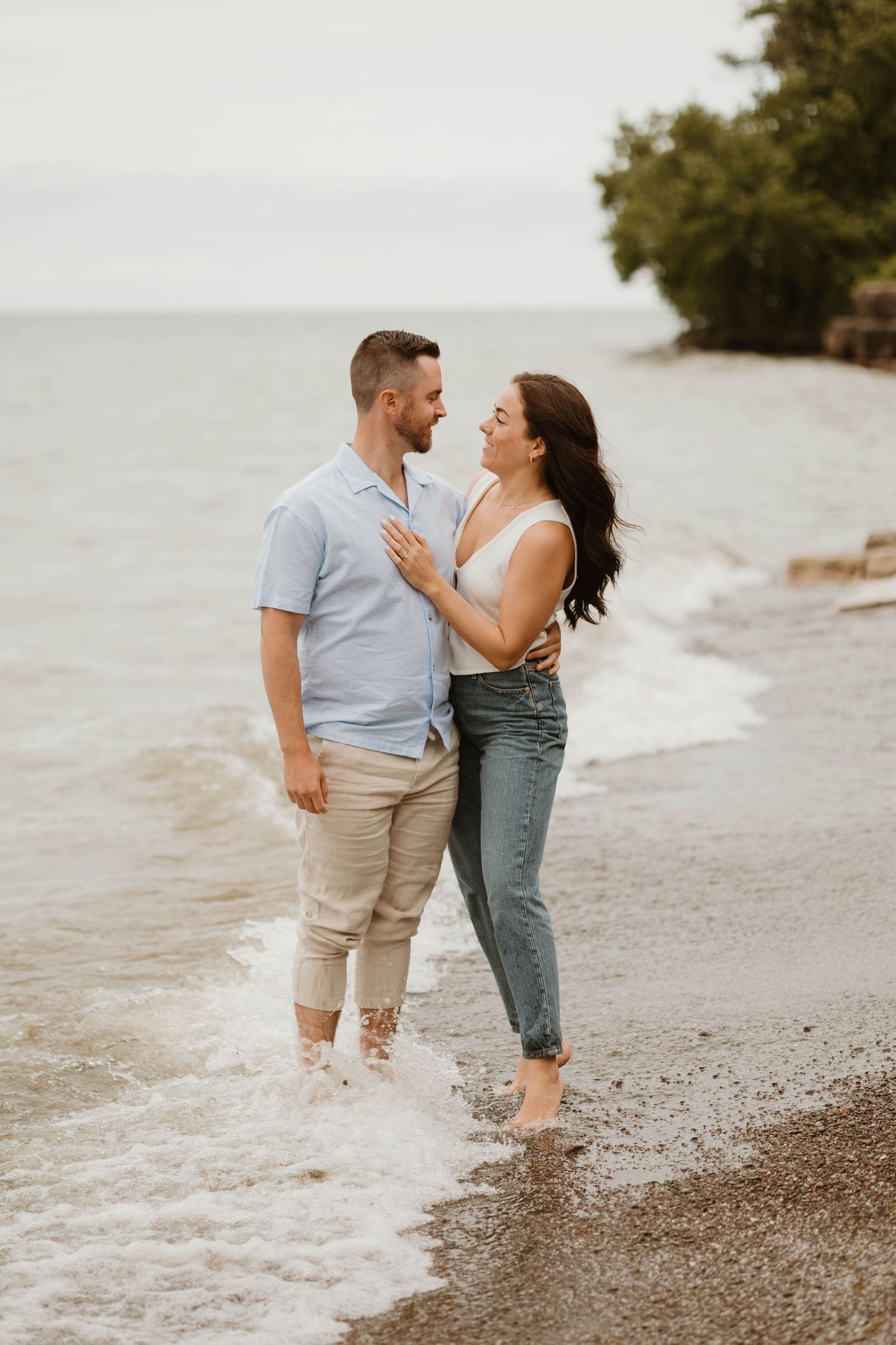 Couple standing in ocean waves on a beach, smiling at each other, woman in jeans and white top.