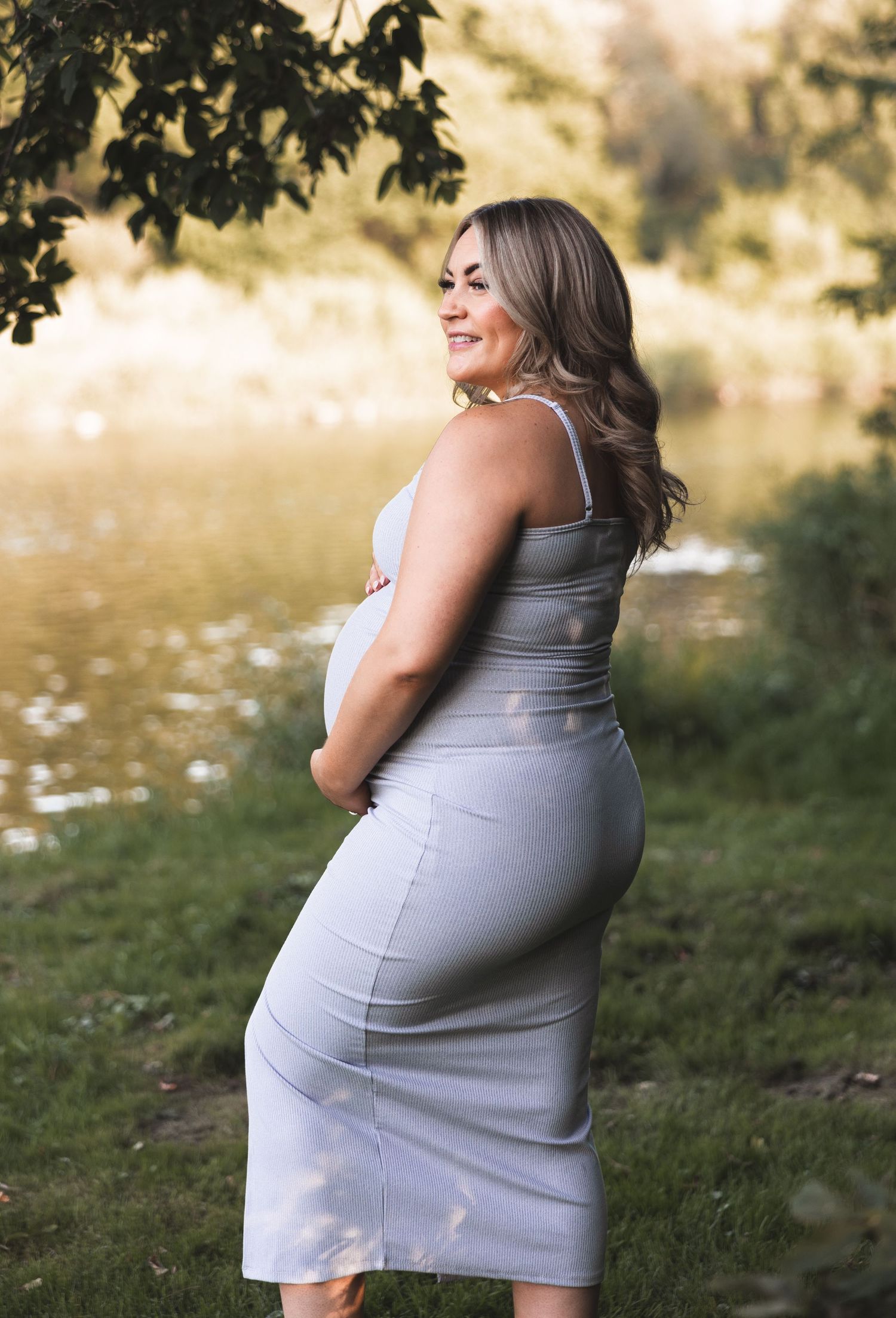 Smiling pregnant woman in gray dress holding baby bump near a pond.