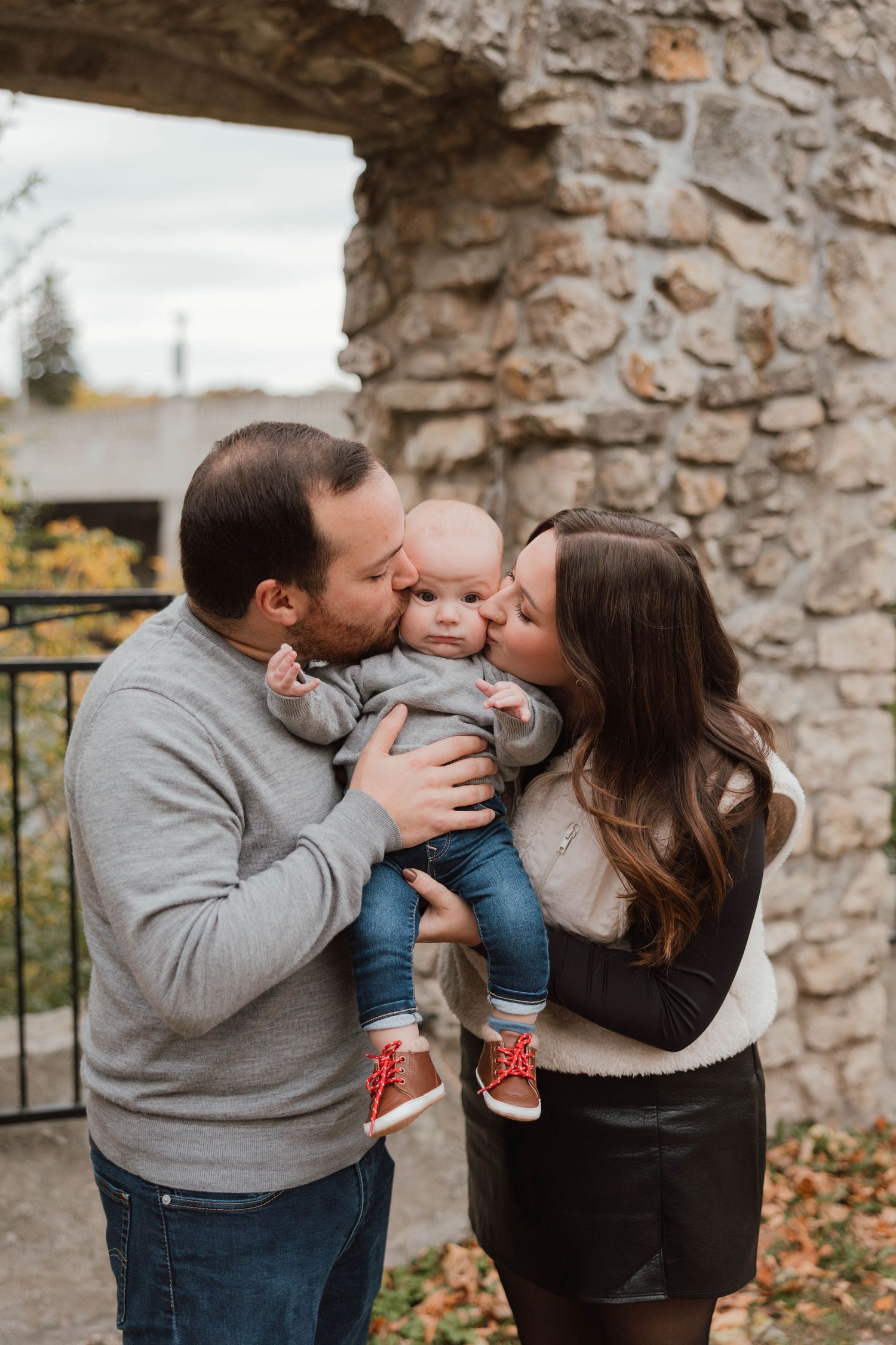 Parents kissing baby on both cheeks simultaneously in front of a rustic stone wall outdoors.