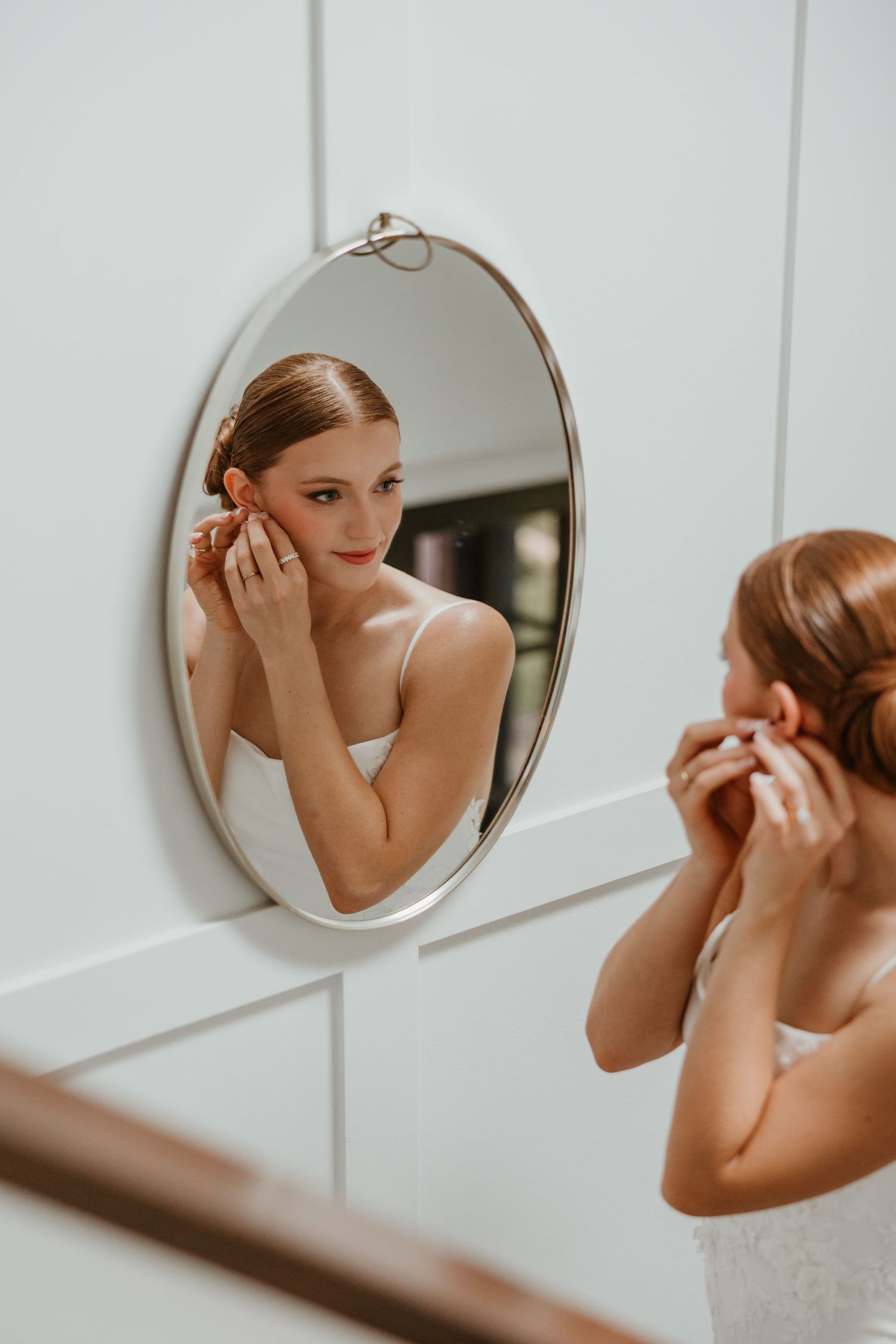 Bride in white strapless gown adjusts pearl earring while looking into an oval mirror.