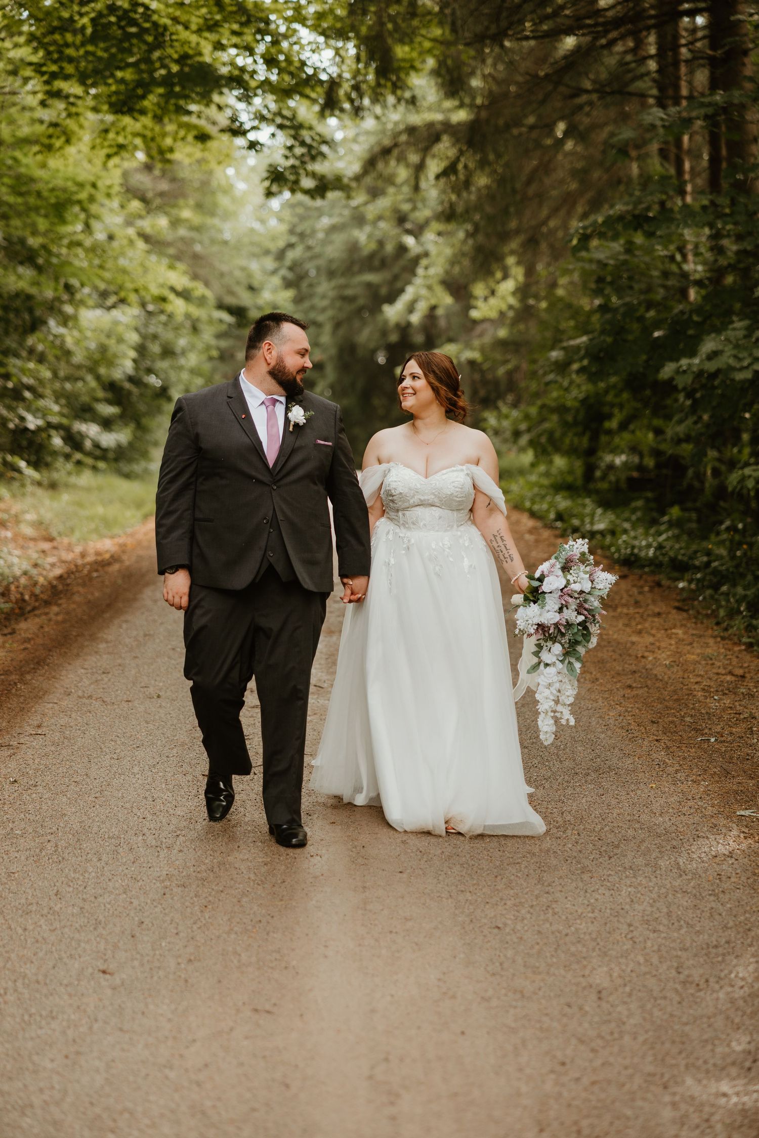 Bride and groom walking hand in hand down a tree-lined path on wedding day.