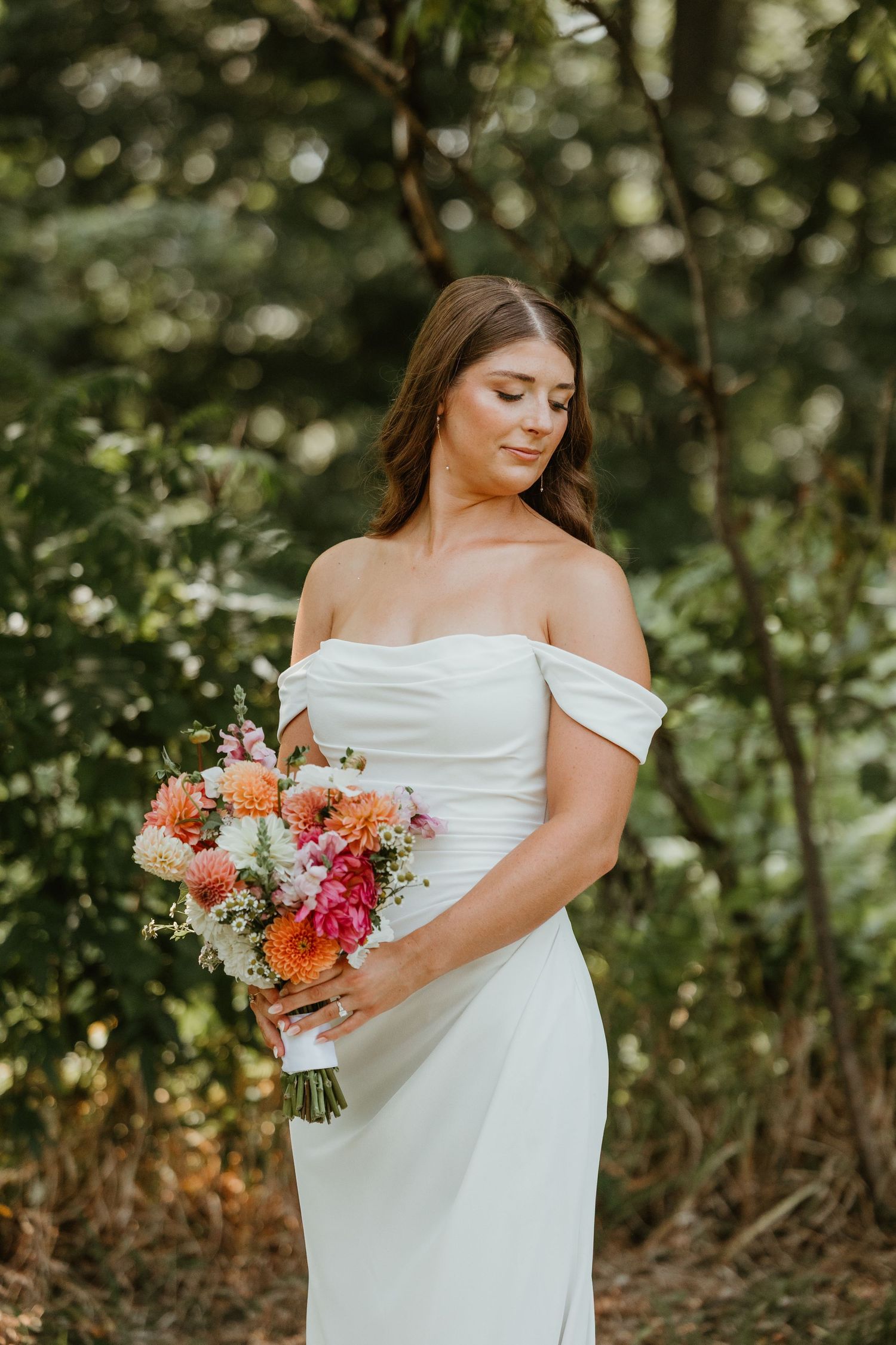 Bride in off-shoulder white gown holding a vibrant pink and orange dahlia bouquet in lush greenery.