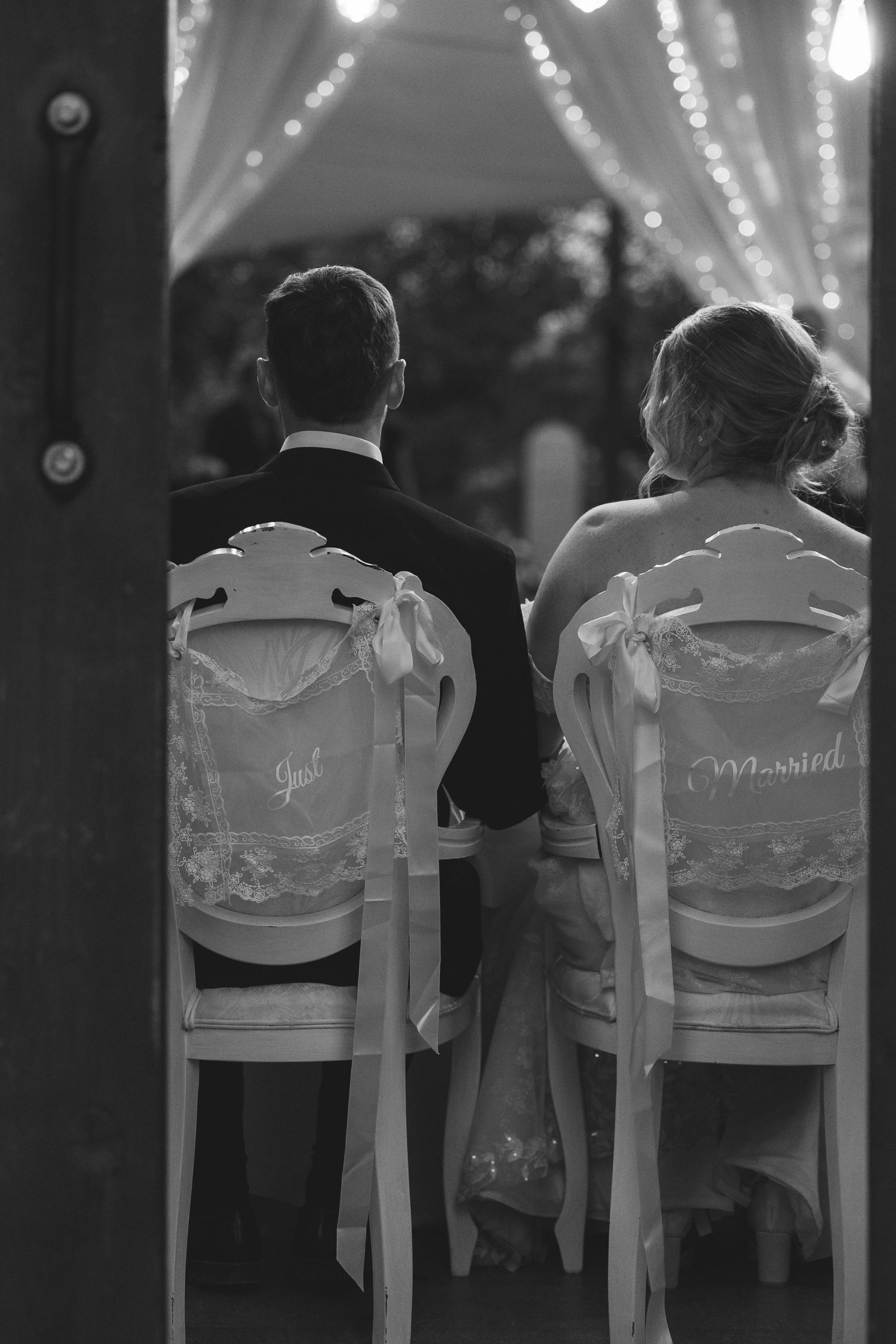 Black and white photo of a bride and groom seated from behind on decorated chairs with Just Married signs under twinkling string lights.