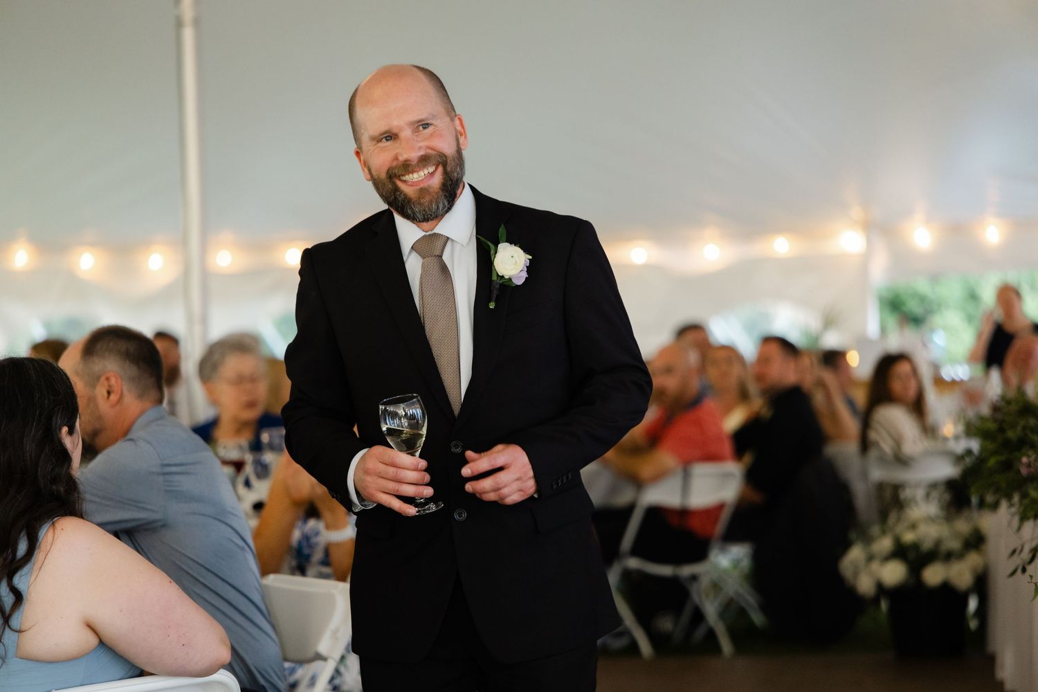 Smiling bald man in black suit holding champagne glass during elegant tented wedding reception.