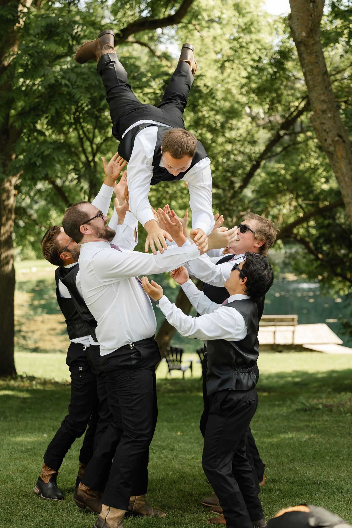 Groomsmen in black vests toss groom into the air outdoors at a wedding celebration on a sunny day.