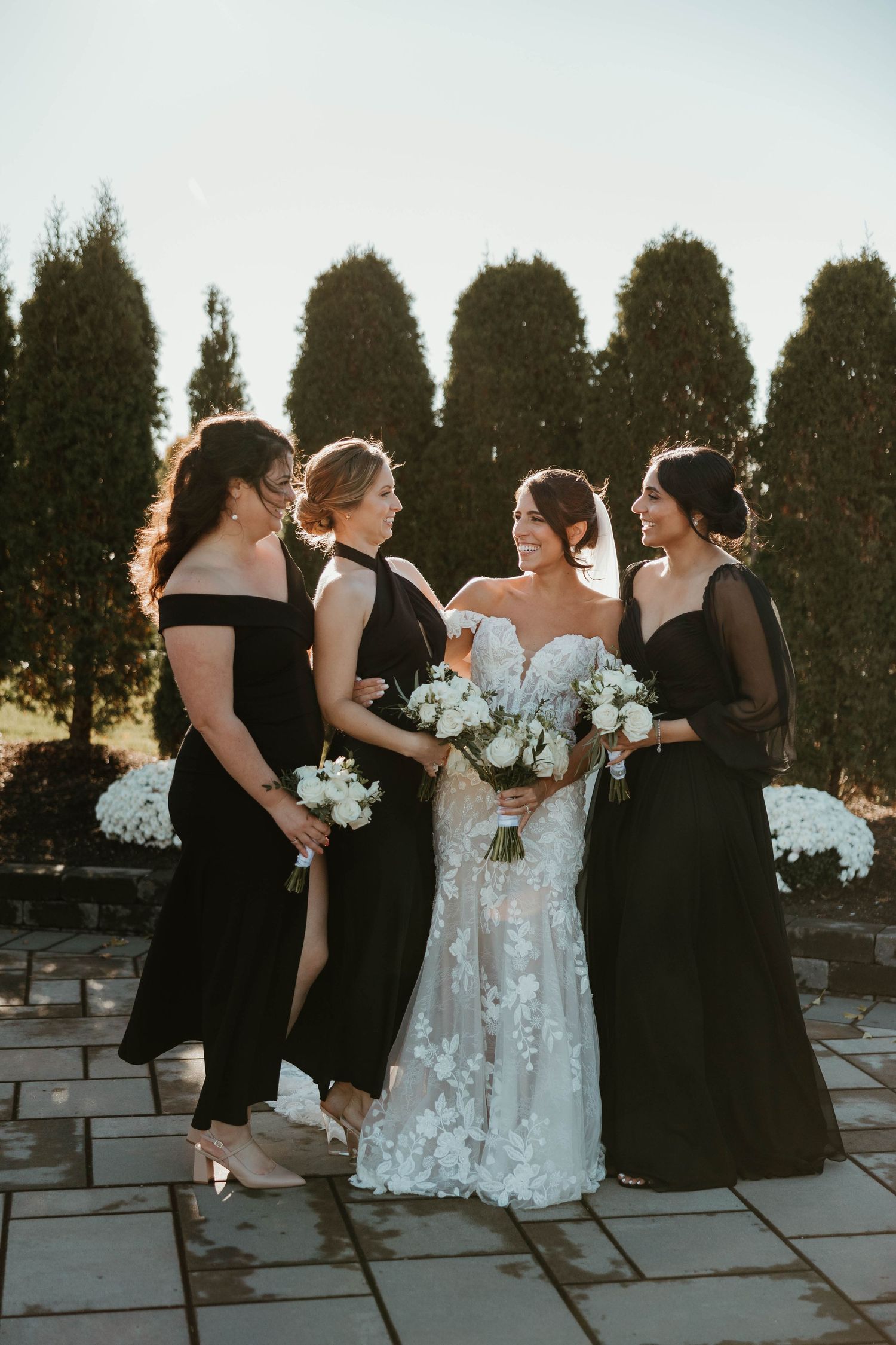 Bride in floral lace gown poses with three bridesmaids in black dresses holding white bouquets.
