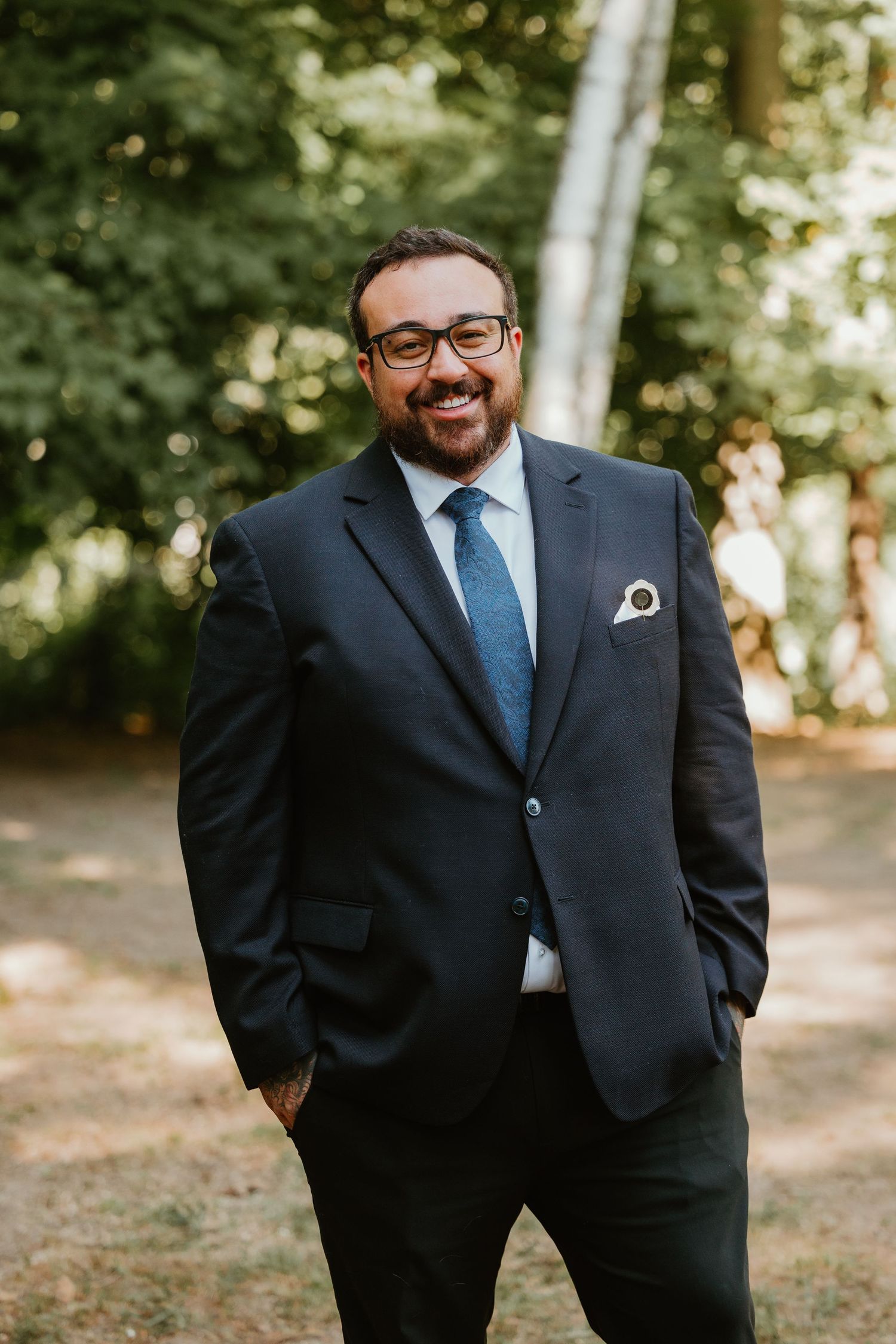 Smiling groom in navy suit with blue tie and pocket square posing outdoors.