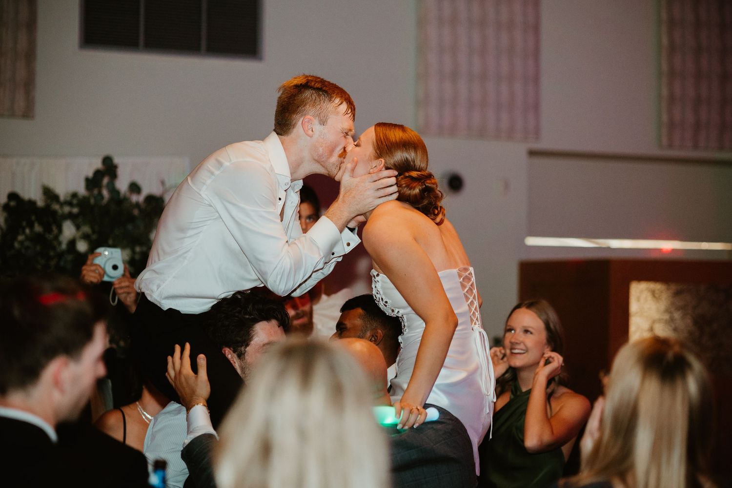 Bride and groom sharing a passionate kiss on the dance floor surrounded by cheering wedding guests.