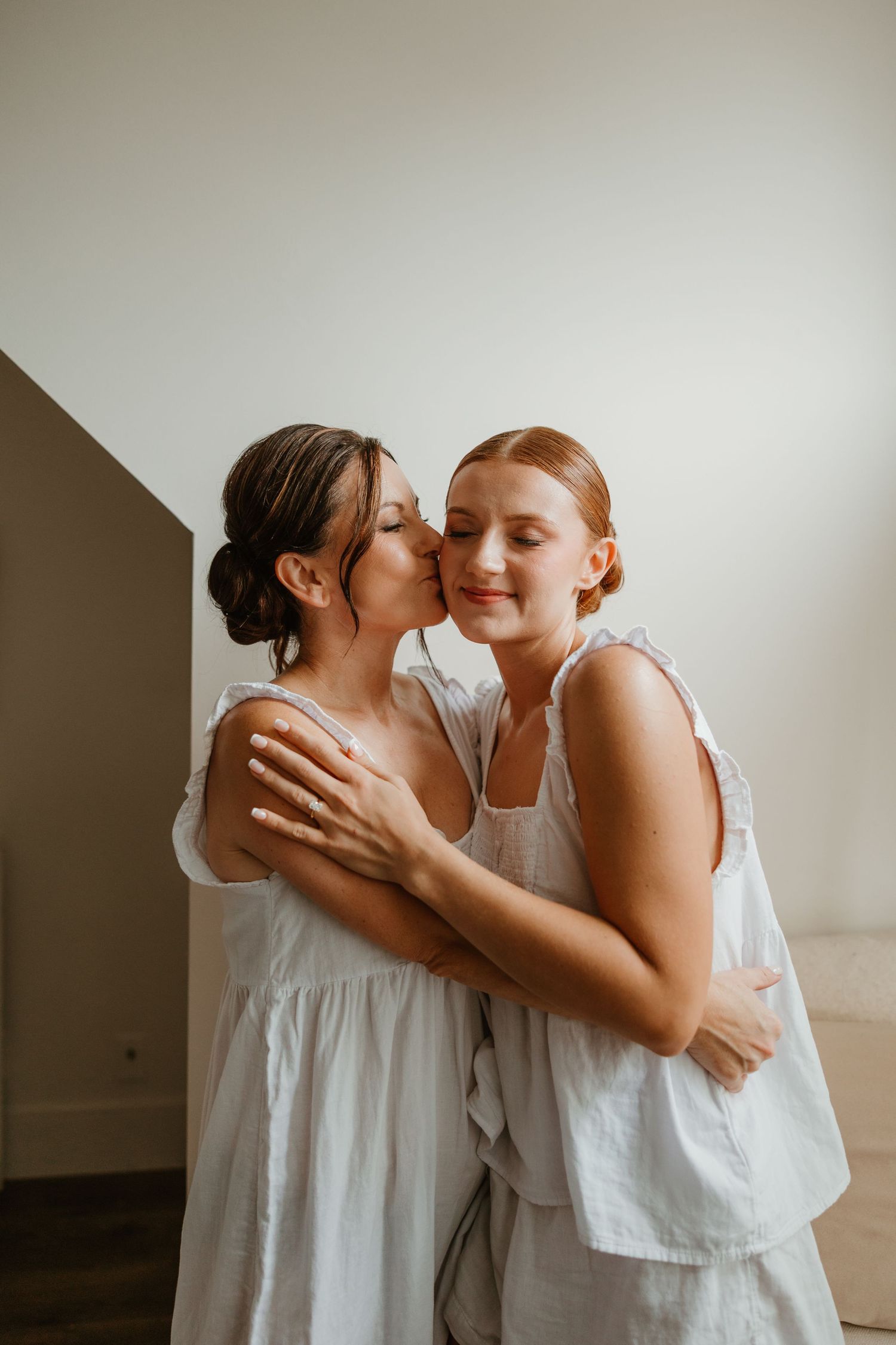 Two women in white dresses share a tender kiss on the cheek during wedding preparations.