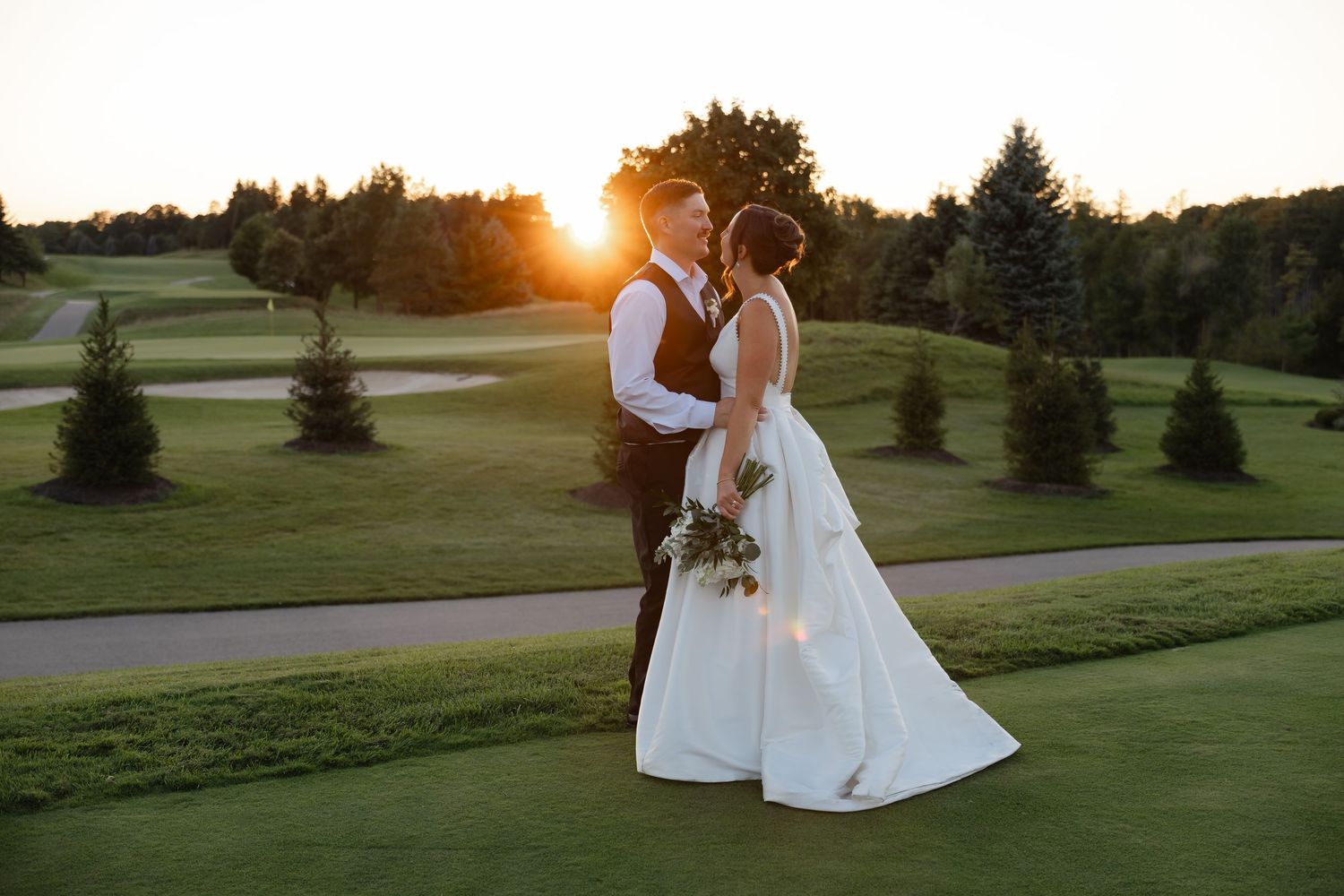 Bride in a white gown and groom in a vest embrace on a golf course fairway at sunset, surrounded by evergreen trees.