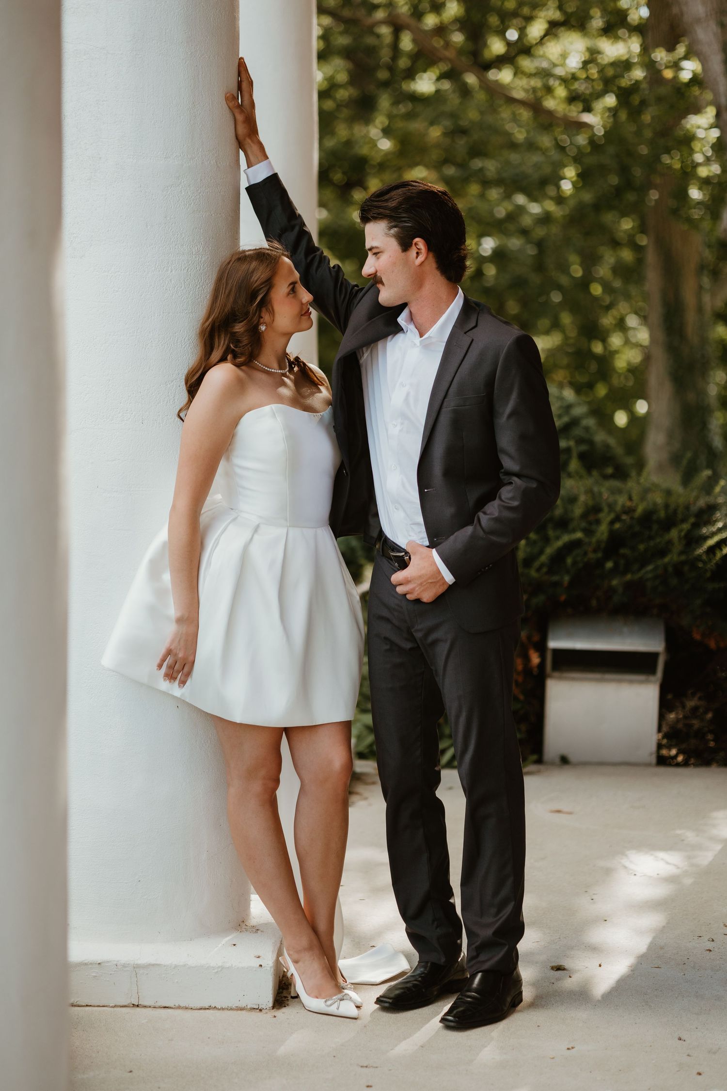 Bride in short white dress and groom in dark suit share a romantic glance between white columns outdoors.