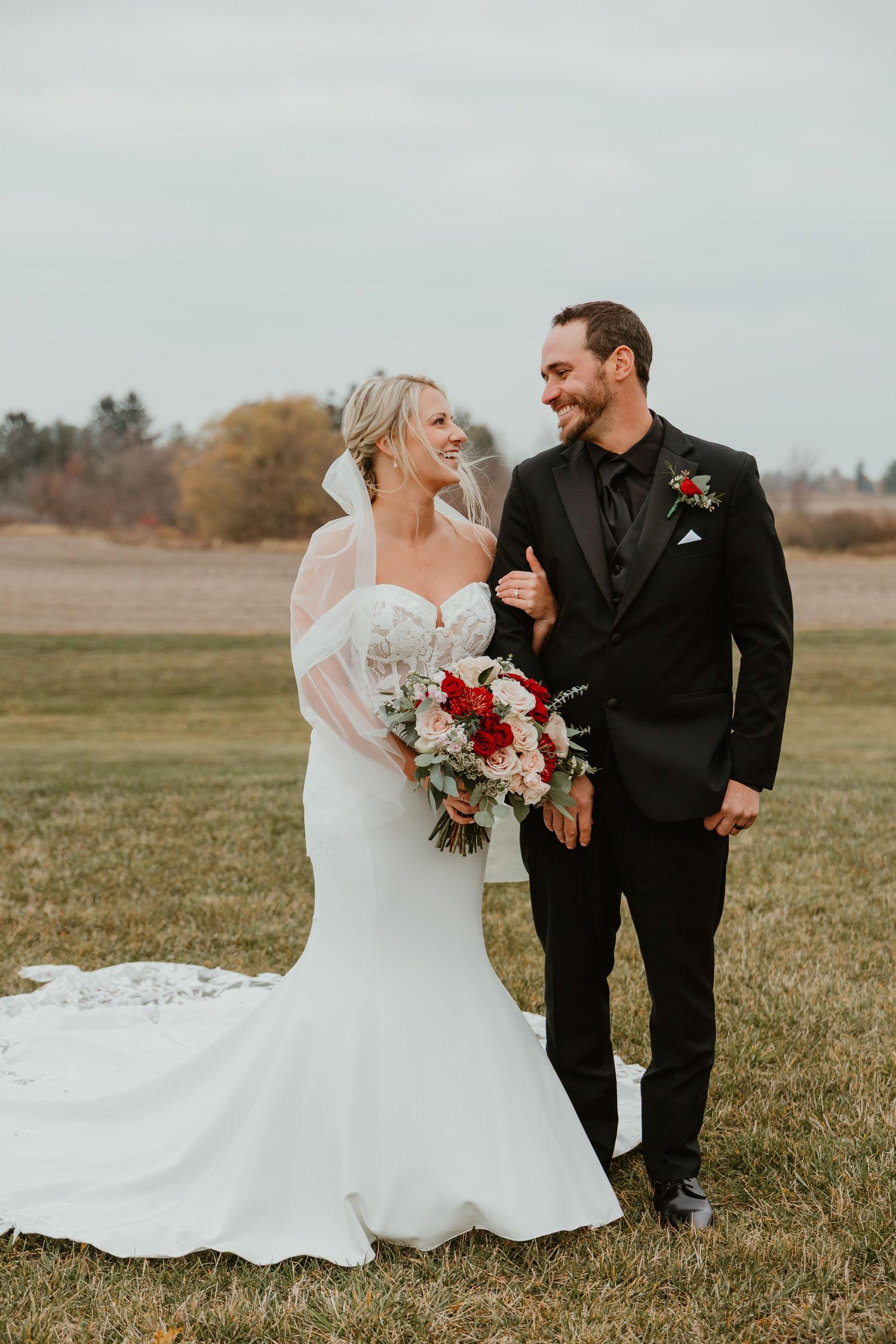 Bride and groom share a joyful moment in a field, bride holding red and pink bouquet in white gown.