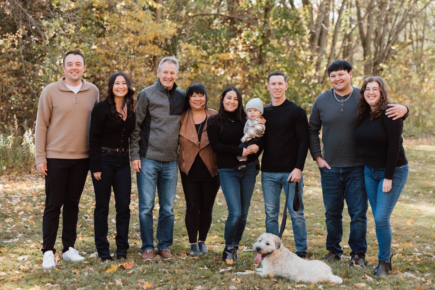 Extended family group of eight adults with a baby and white dog posing outdoors in autumn foliage.