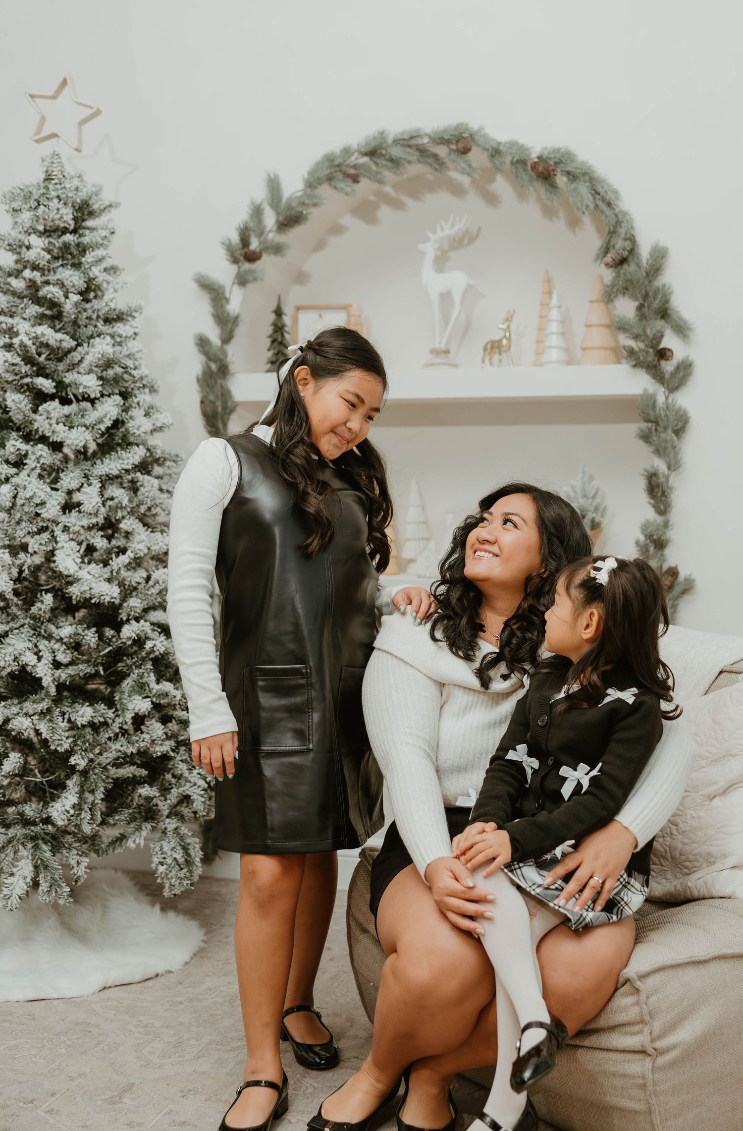Mother sitting with two daughters near flocked Christmas tree in festive holiday setting.