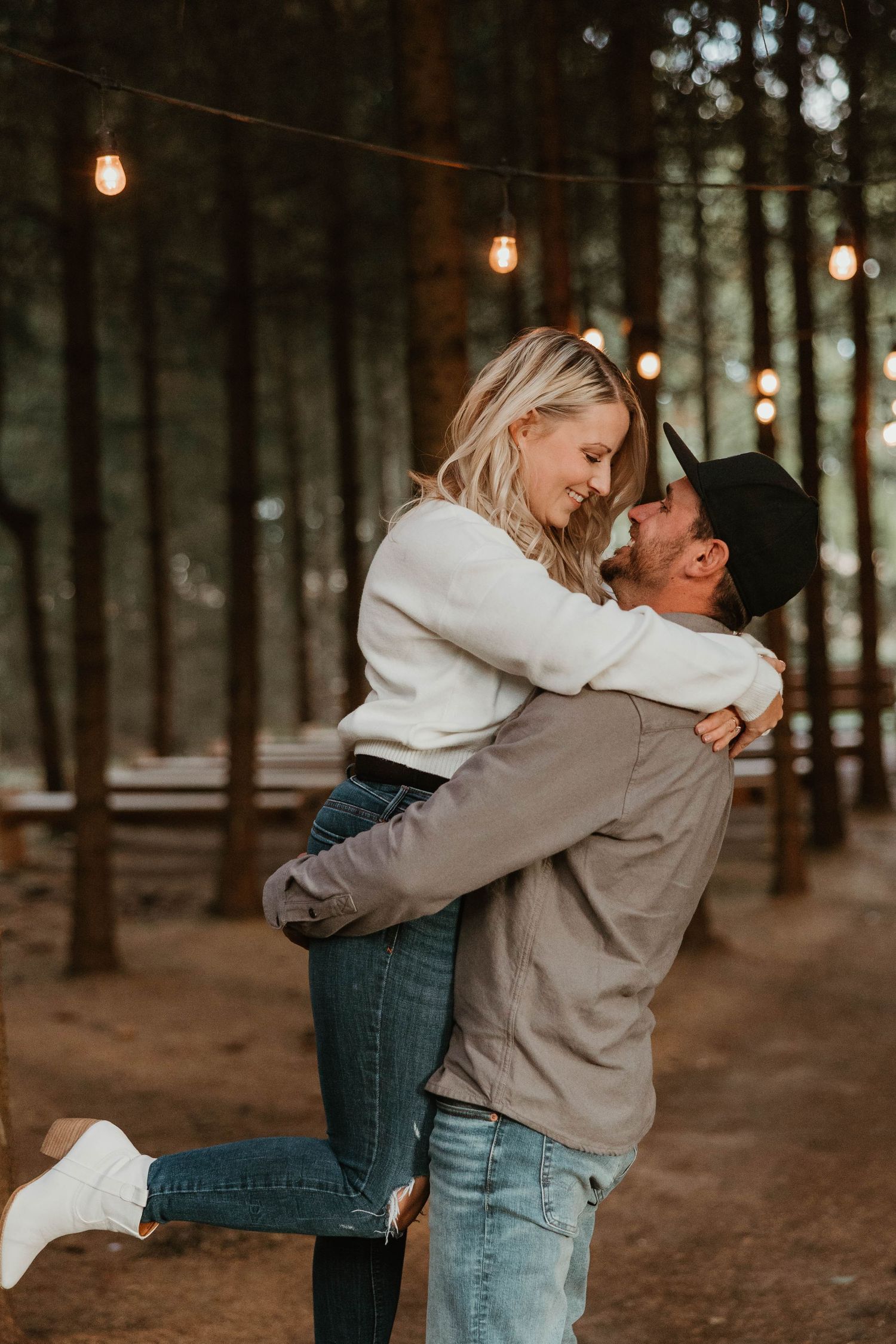 Man lifting woman in white sweater and jeans in pine forest with string lights.