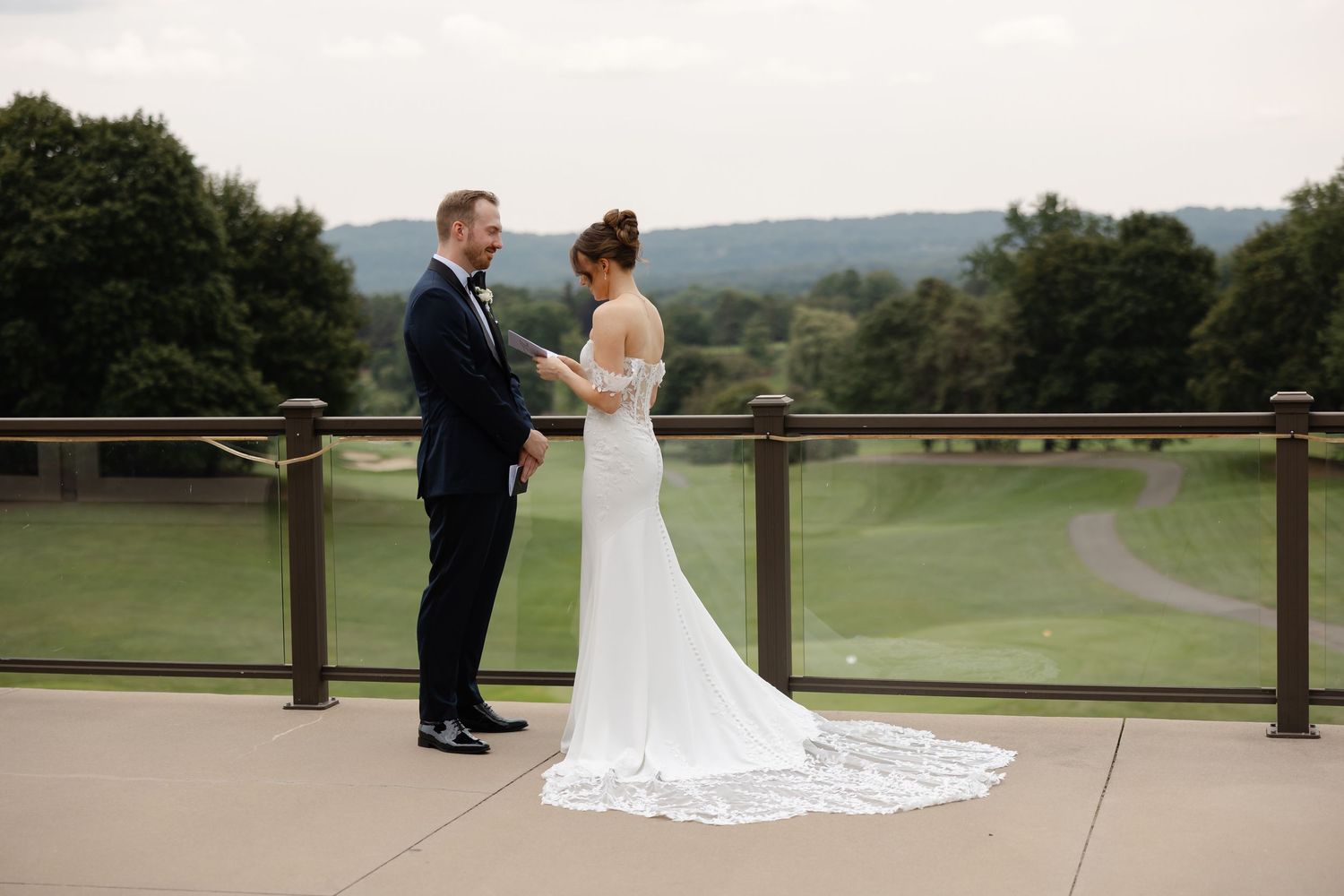 Bride and groom share first look on outdoor terrace with scenic golf course and rolling hills backdrop.