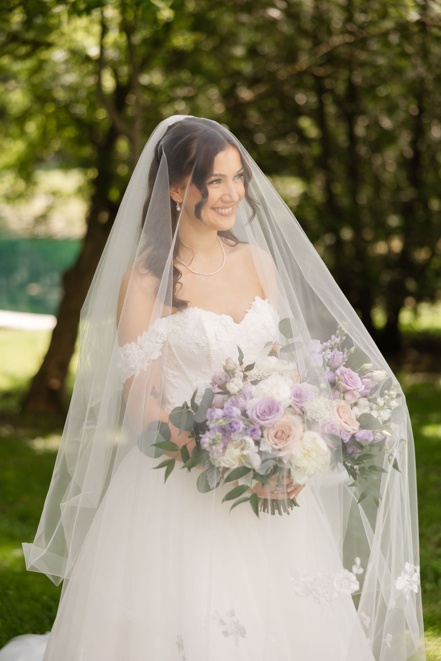 Elegant bride in strapless white gown and veil holds lavender and blush bouquet outdoors among green trees.