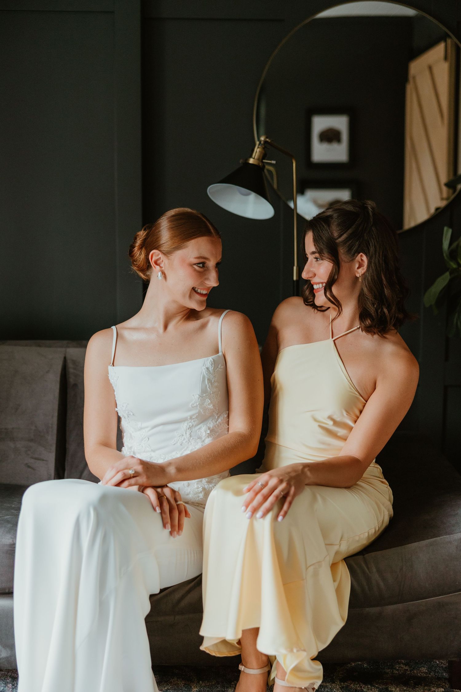 Bride in white dress and bridesmaid in yellow dress sitting together on a gray sofa smiling at each other.