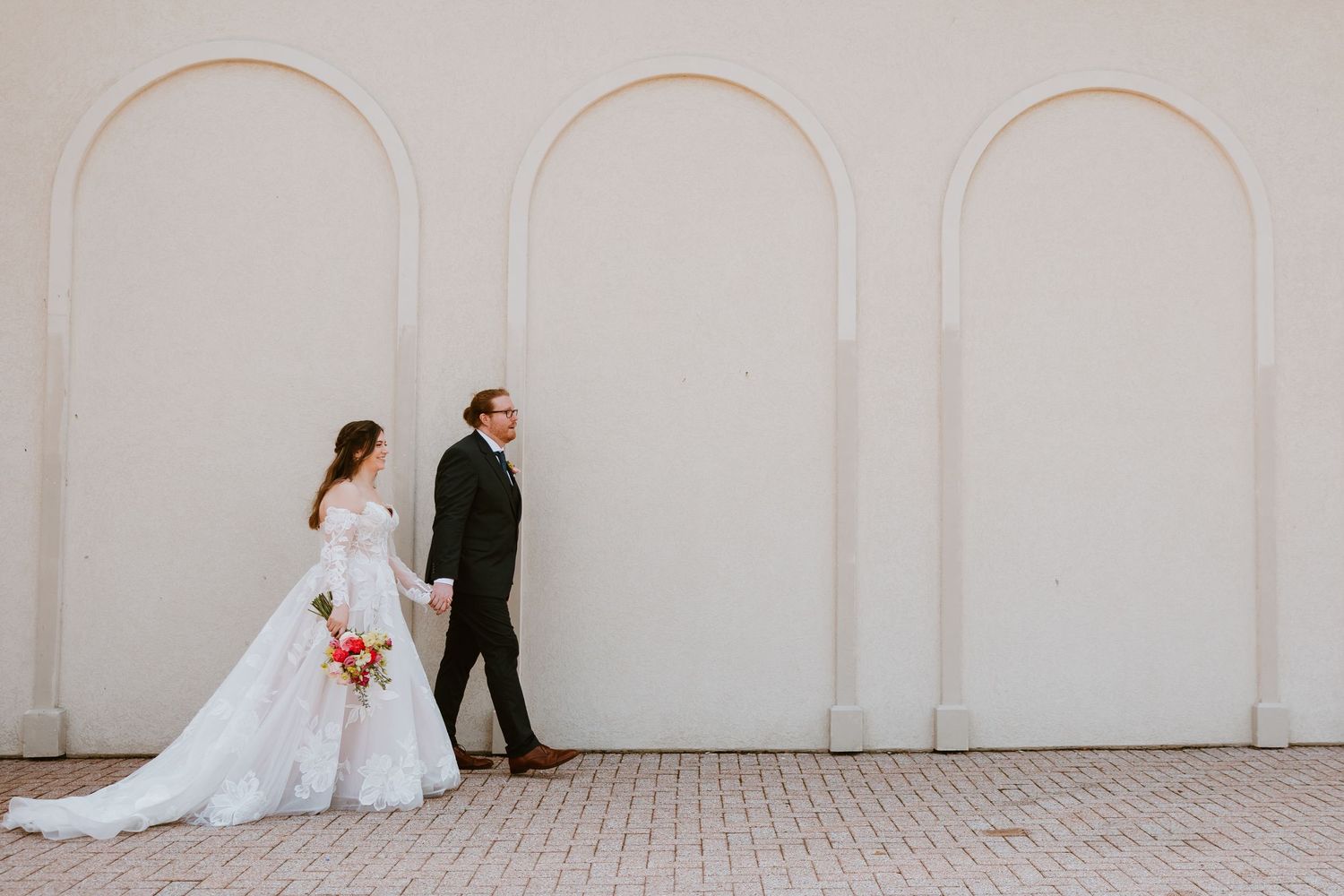 Bride in white gown and groom in black suit walk hand in hand beside elegant arched white wall on wedding day.
