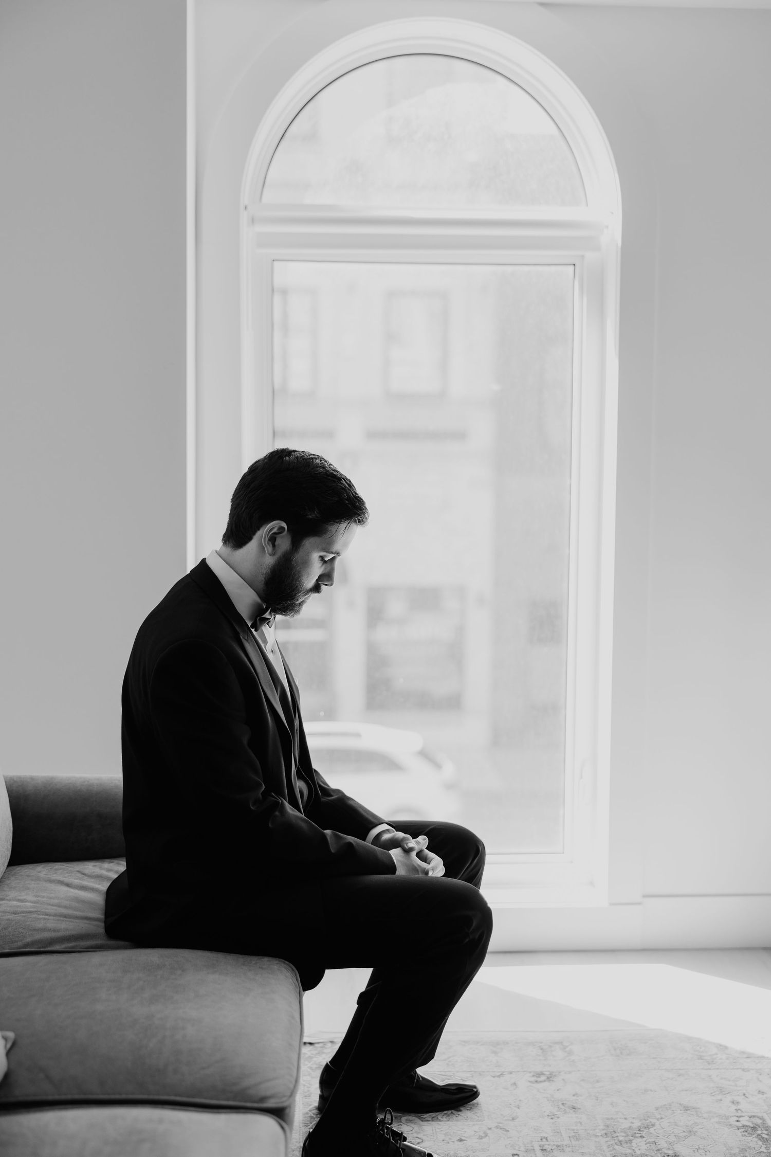 Black and white portrait of a bearded groom sitting on a sofa by an arched window before his wedding.