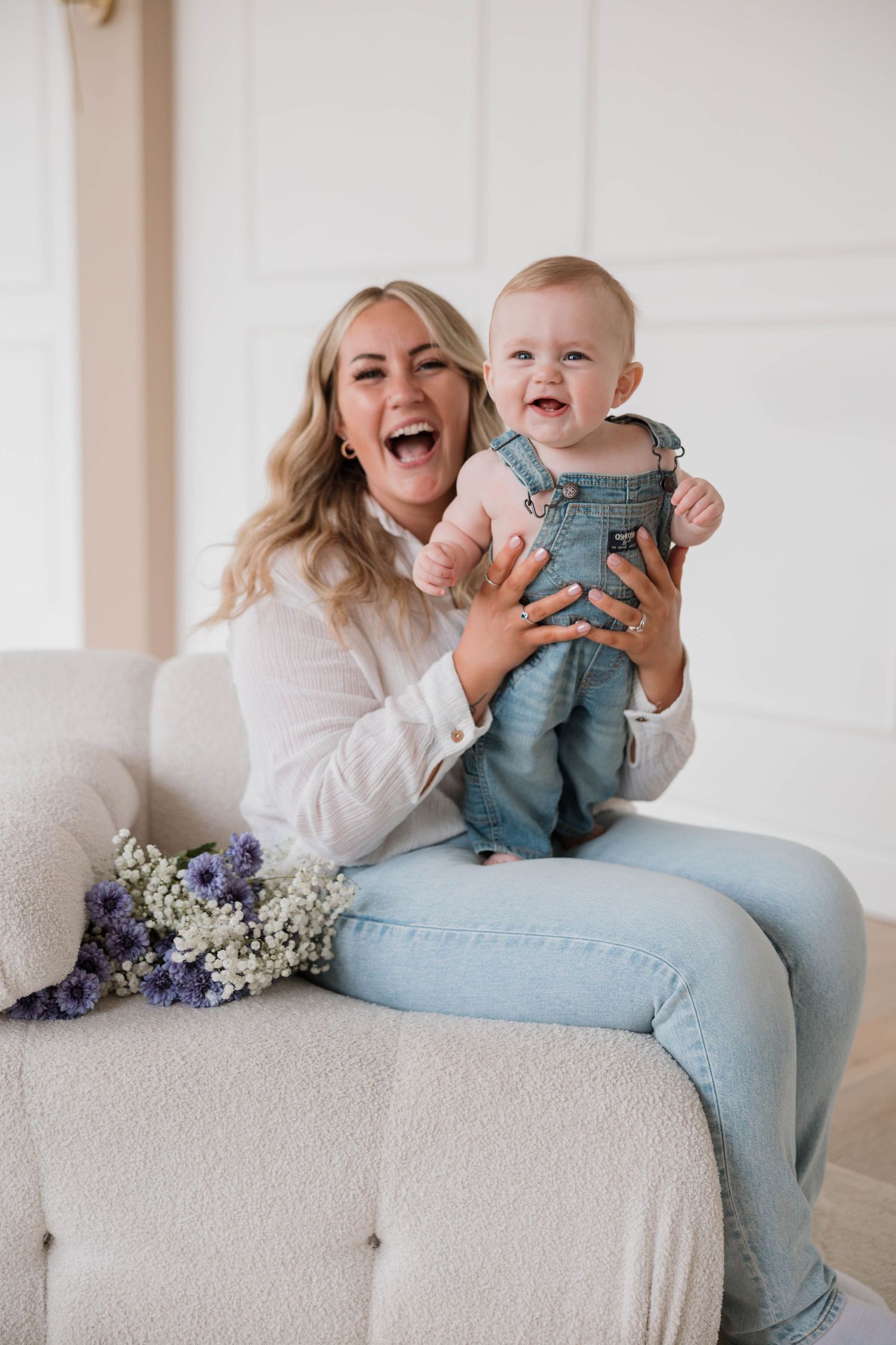 Laughing mother holding smiling baby in denim overalls on white sofa with purple flowers.