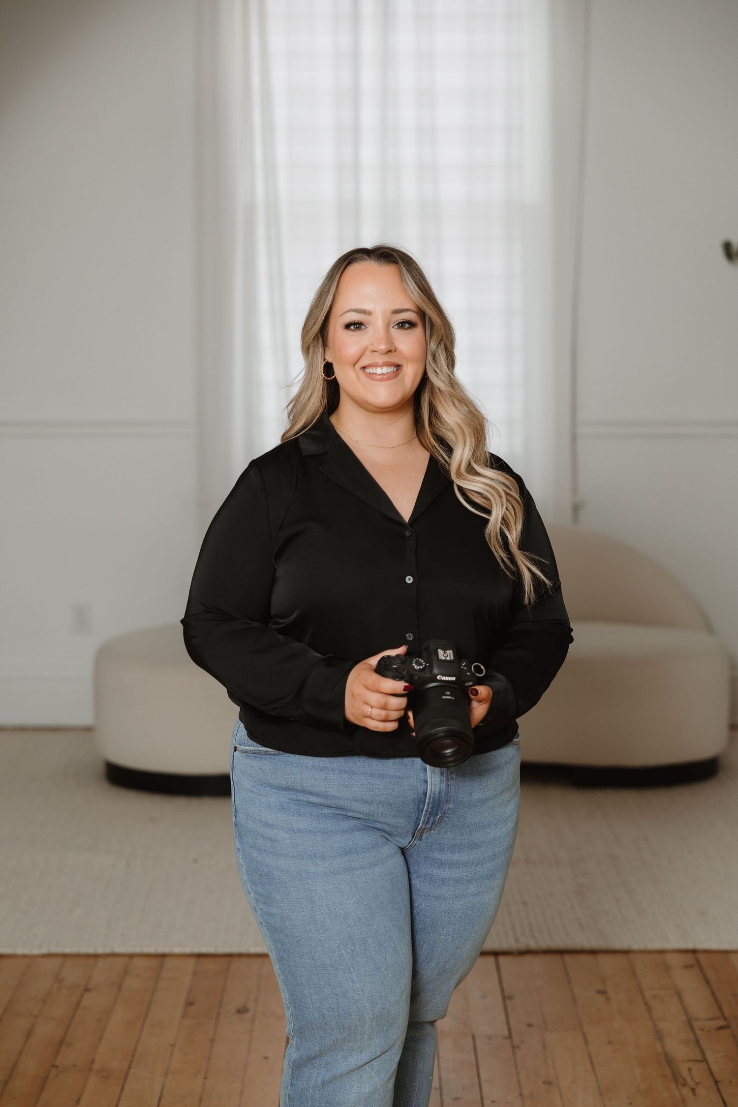 Female photographer smiling and holding camera in bright studio wearing black shirt and jeans.
