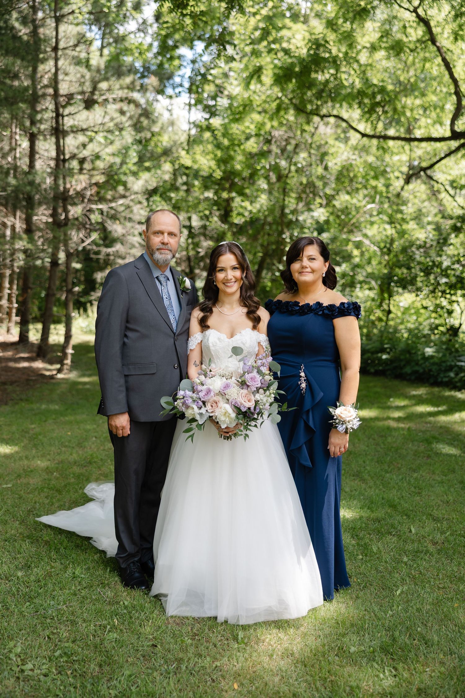 Bride in white ball gown holding floral bouquet poses with parents on green lawn surrounded by tall trees.